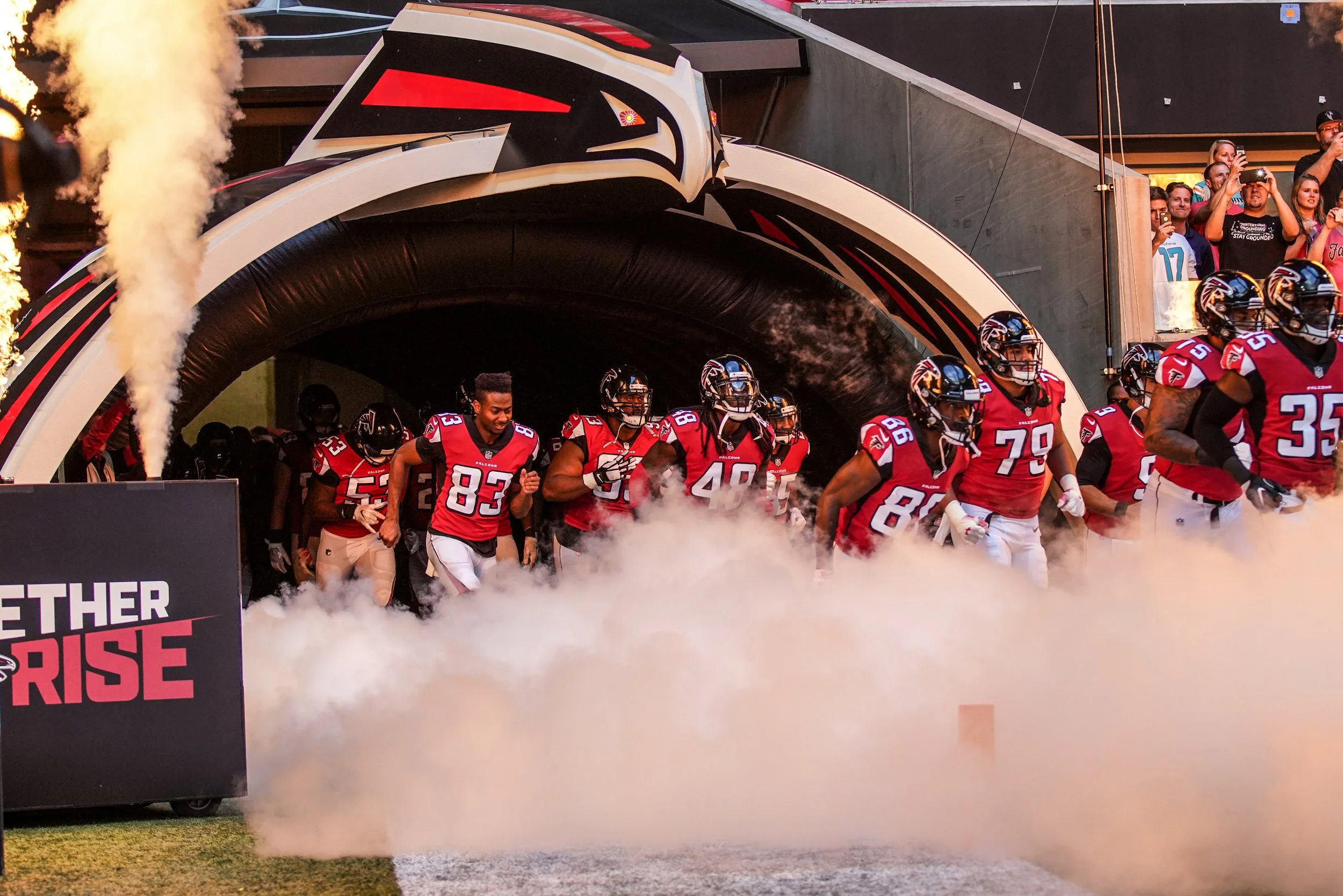 Football team in red uniforms running out of tunnel onto field, with smoke and steam effects, spectators watching from stands