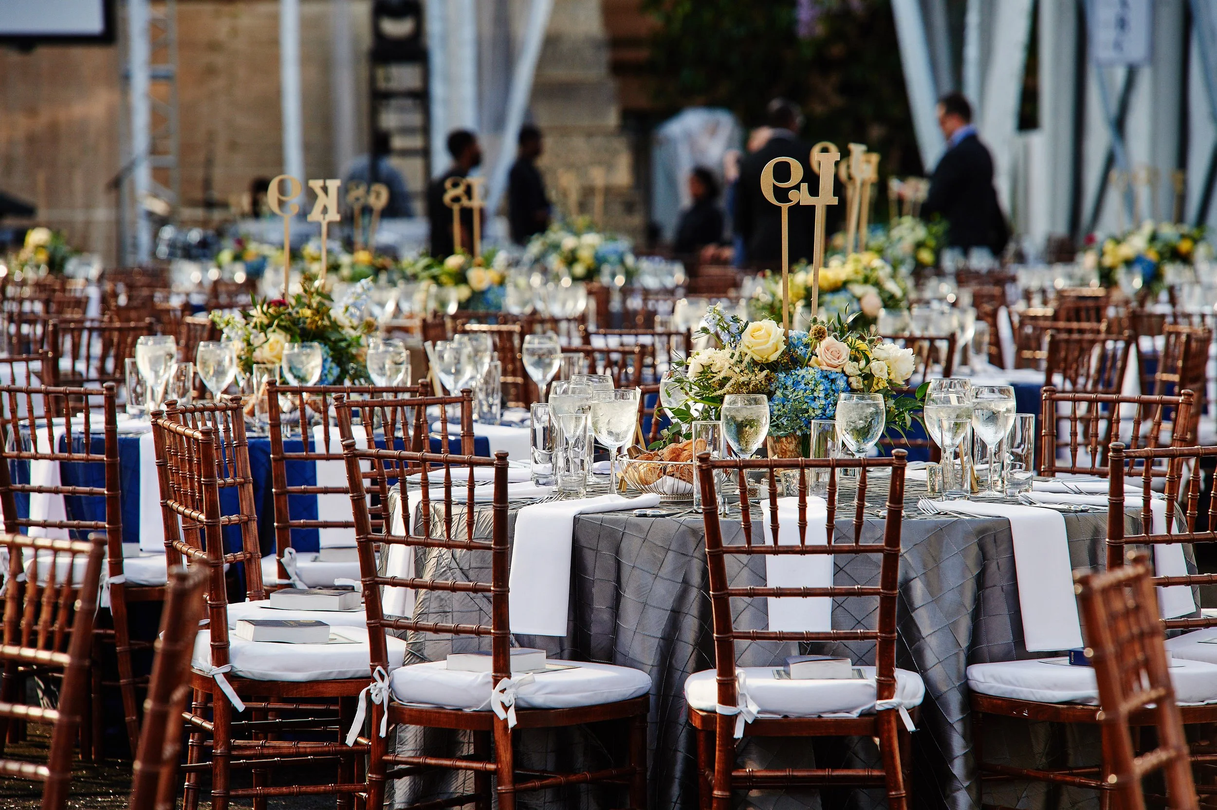 Arranged outdoor banquet tables with floral centerpieces, glassware, and chairs, set up for a formal event.