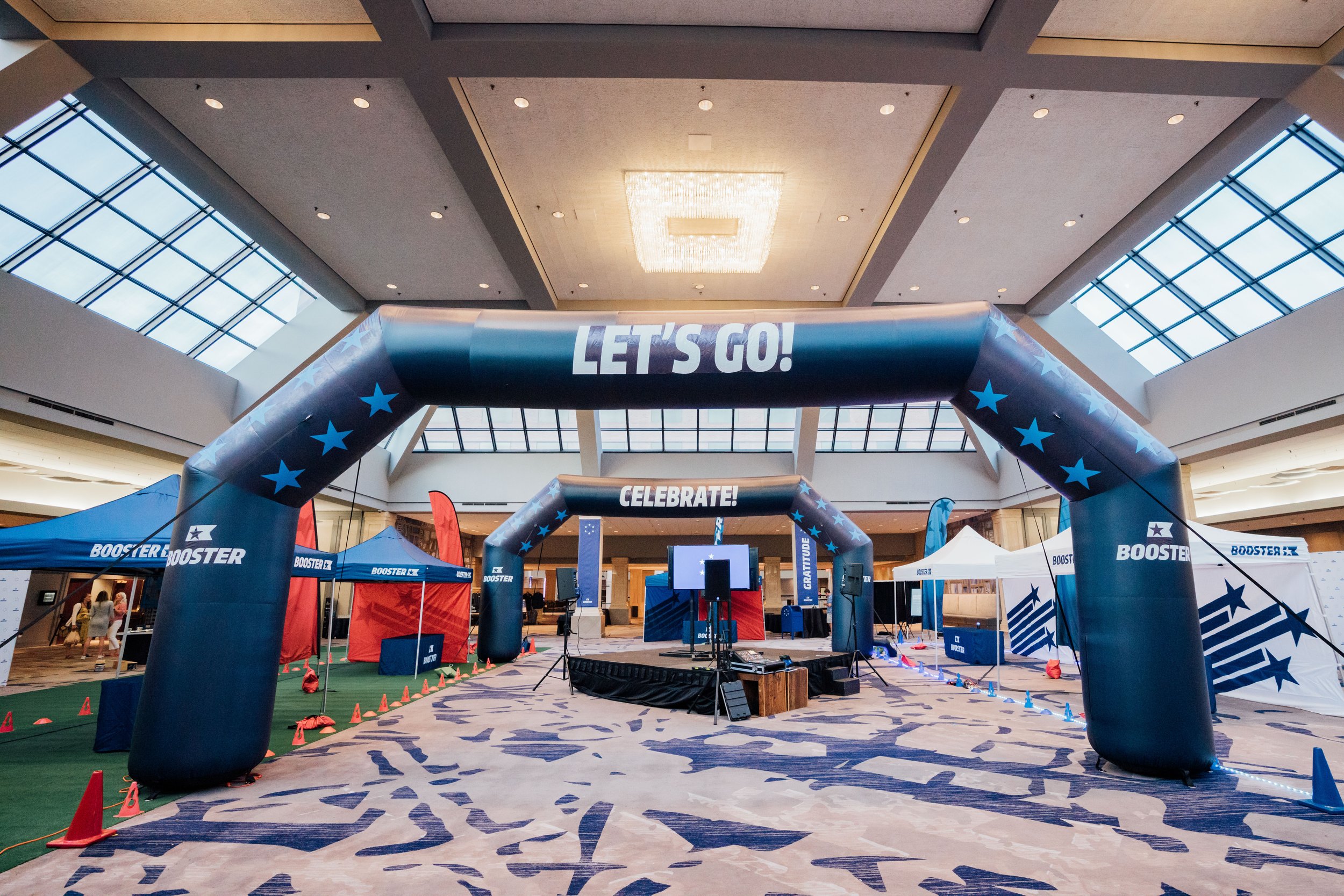 Indoor event space with two large blue inflatable arches that have stars and the words 'LET'S GO!' and 'CELEBRATE!' on them. There are tents, equipment, and a stage with a screen at the center.