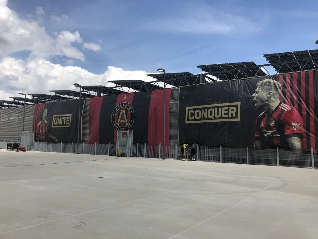 Large banners at Atlanta United FC stadium featuring a female player and a male player in red and black soccer jerseys, with the team's logo and the words 'Unite' and 'Conquer'.