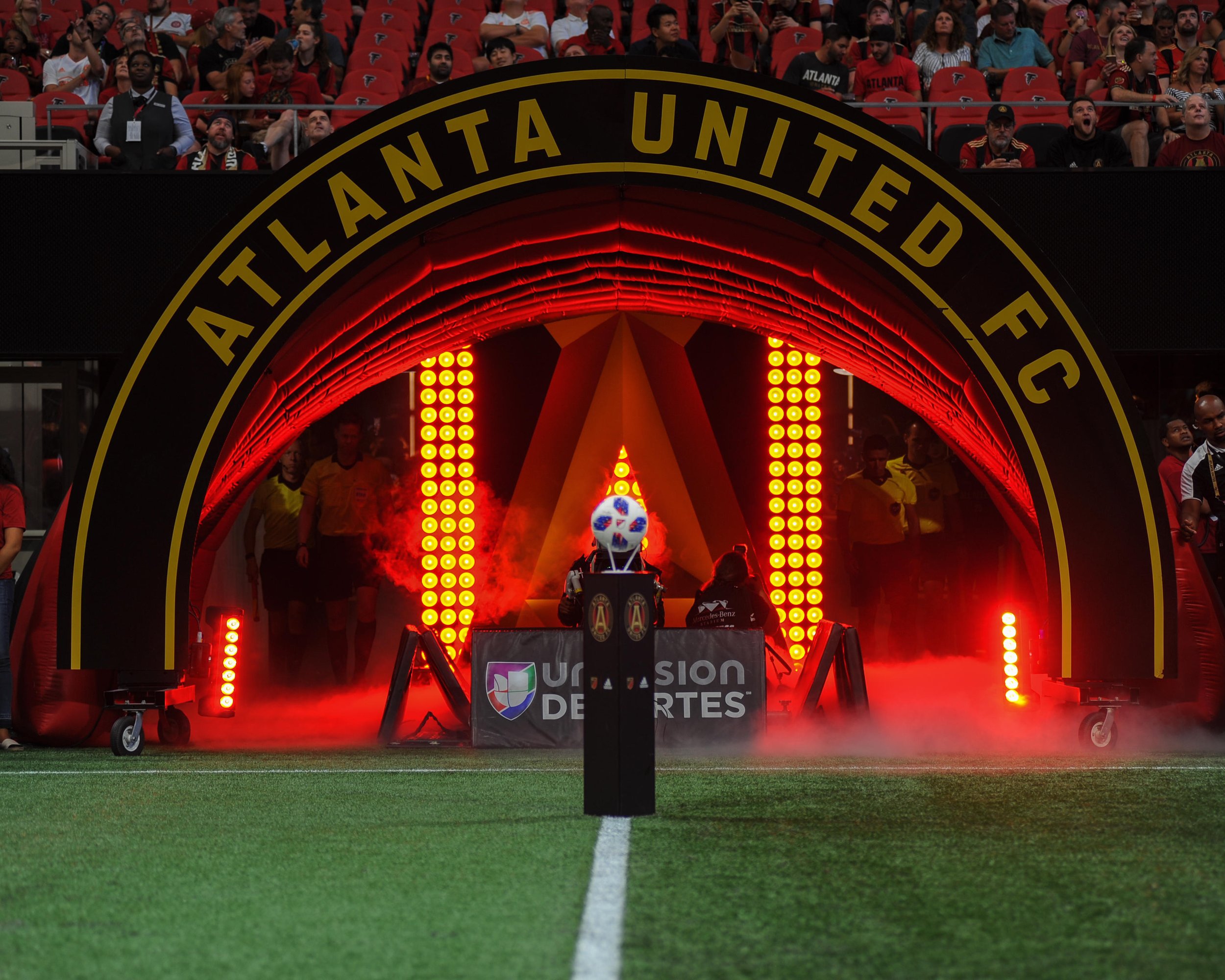 Entrance tunnel at Atlanta United FC soccer stadium with bright red lights, fog, a soccer ball, and spectators in the background.
