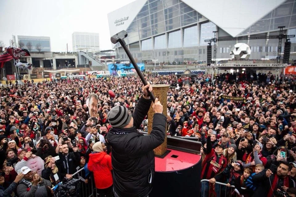 A person holding a hammer stands on a stage with a microphone, addressing a large crowd at an outdoor event with modern buildings in the background.