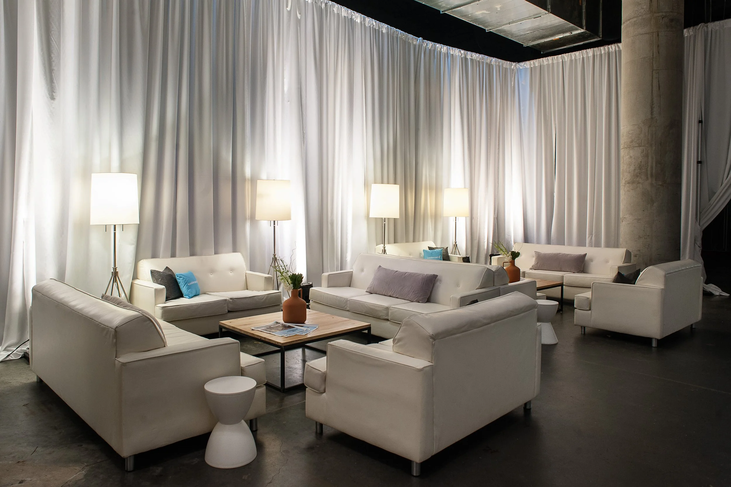 A spacious lounge area with white sofas, side tables, tall lamps, and decorative vases, set against white curtains in an indoor space.