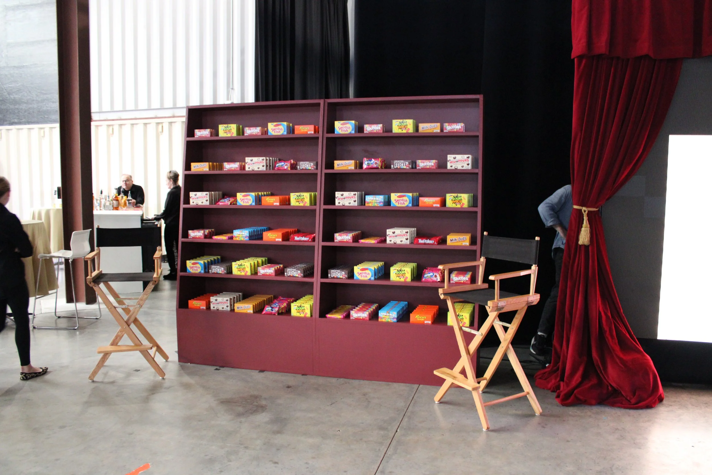 A bookshelf with various candy boxes, two director's chairs on either side, a velvet curtain on the right, and people in the background, likely in a theater or event space.