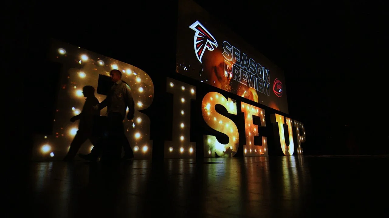 Illuminated letters spelling 'RISE UP' with two people walking past, and a screen displaying 'SEASON PREVIEW' with the Atlanta Falcons logo.