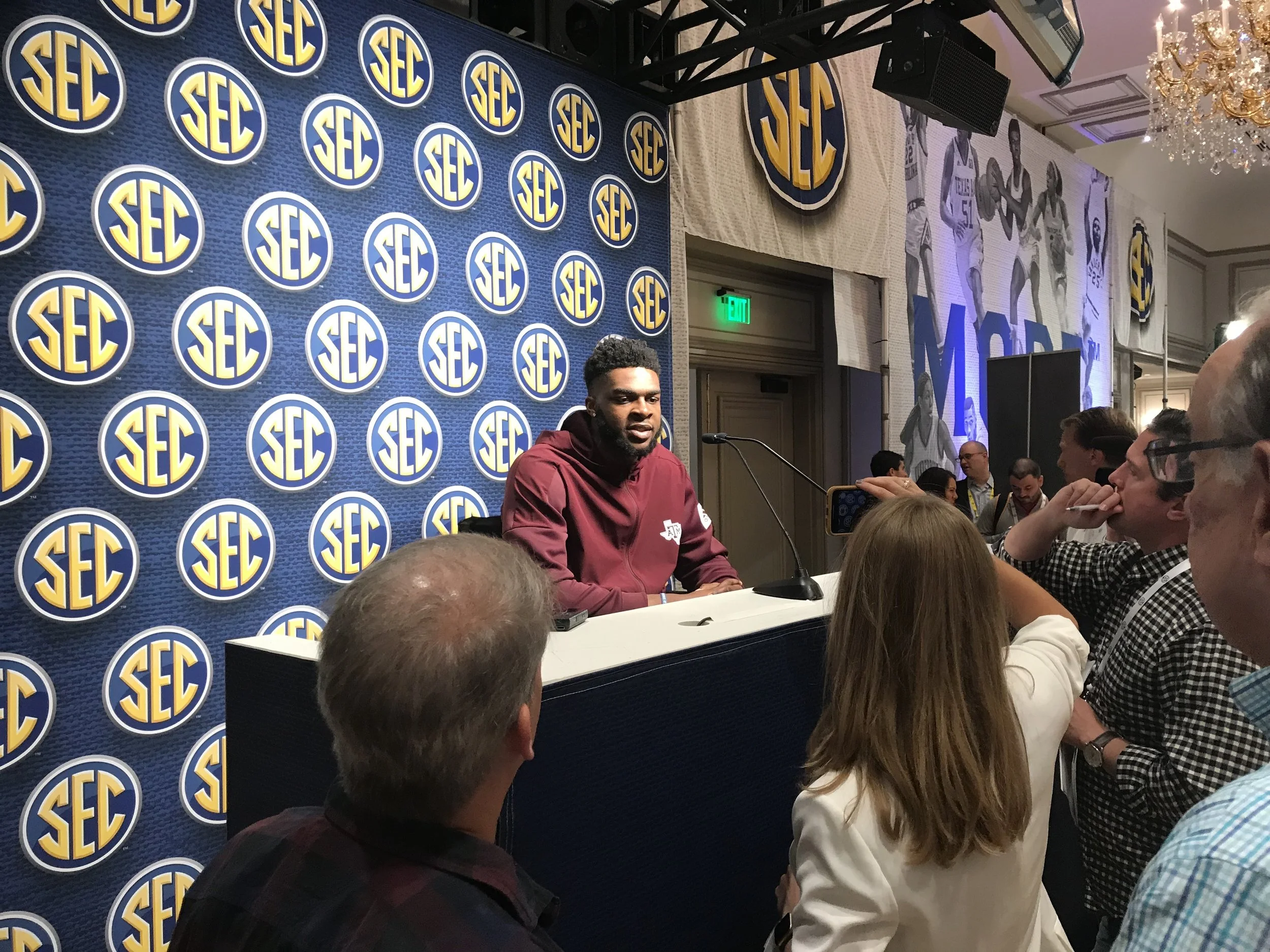 A man wearing a red hoodie speaking at a press conference with an SEC backdrop, surrounded by media and attendees.