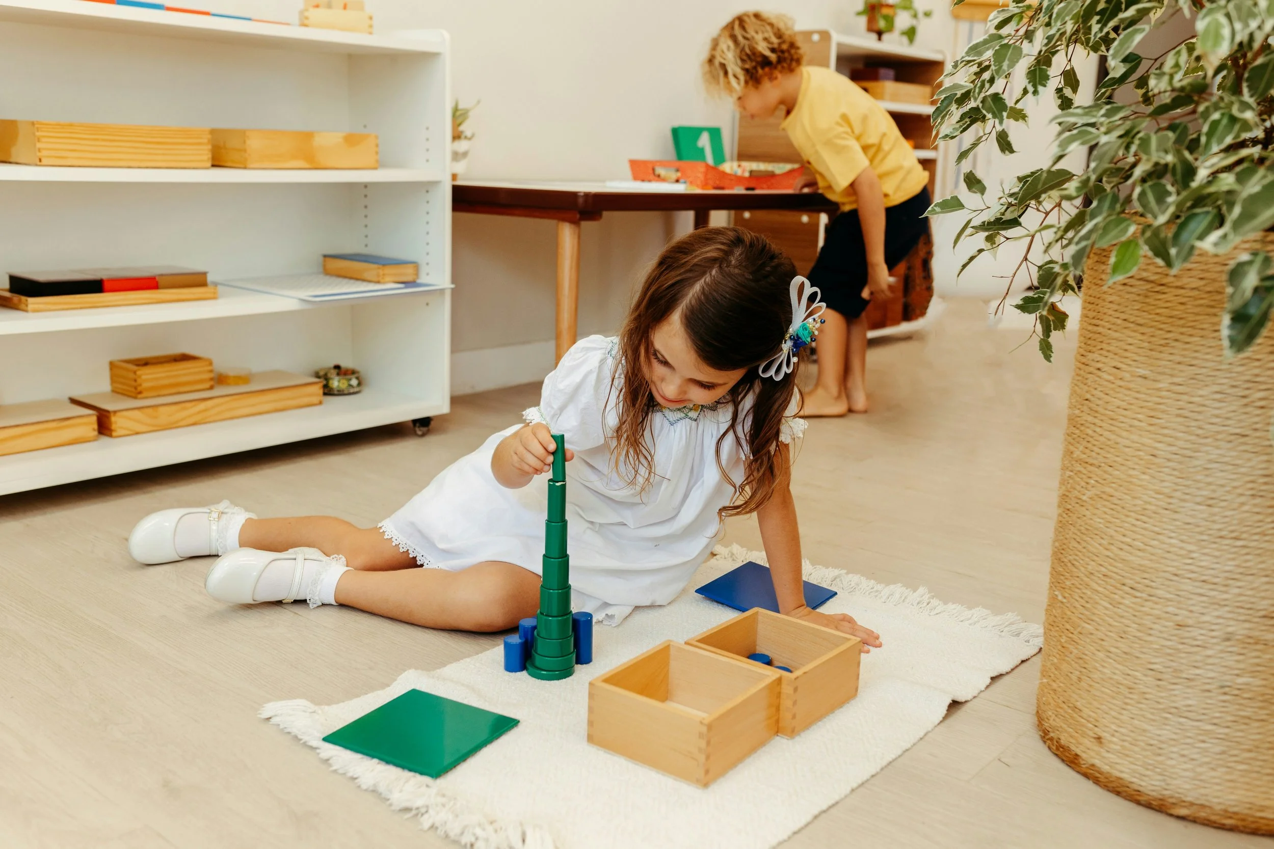 Zwei Kinder spielen auf einem Teppichboden, eins sitzt und baut einen Turm aus grünen Bauklötzen, das andere steht im Hintergrund an einem Tisch.