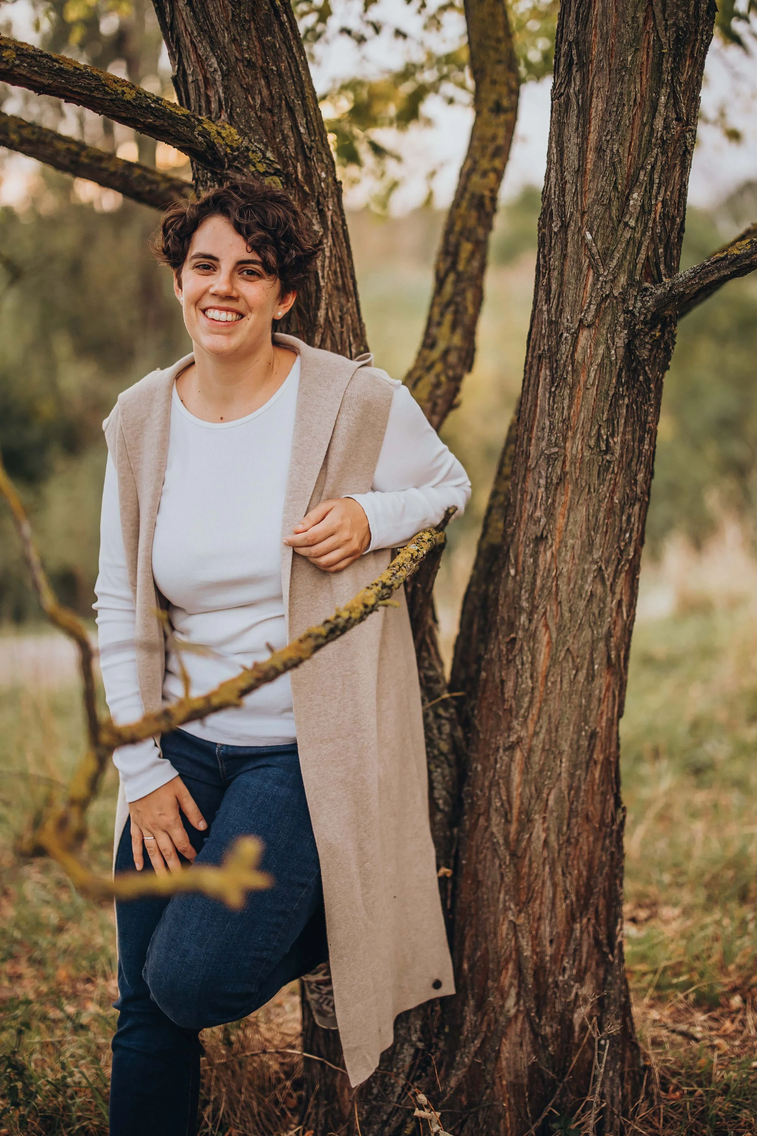 Alexandra Stagl steht draußen an einem Baum im herbstlichen Park, trägt eine weiße Bluse und Jeans.