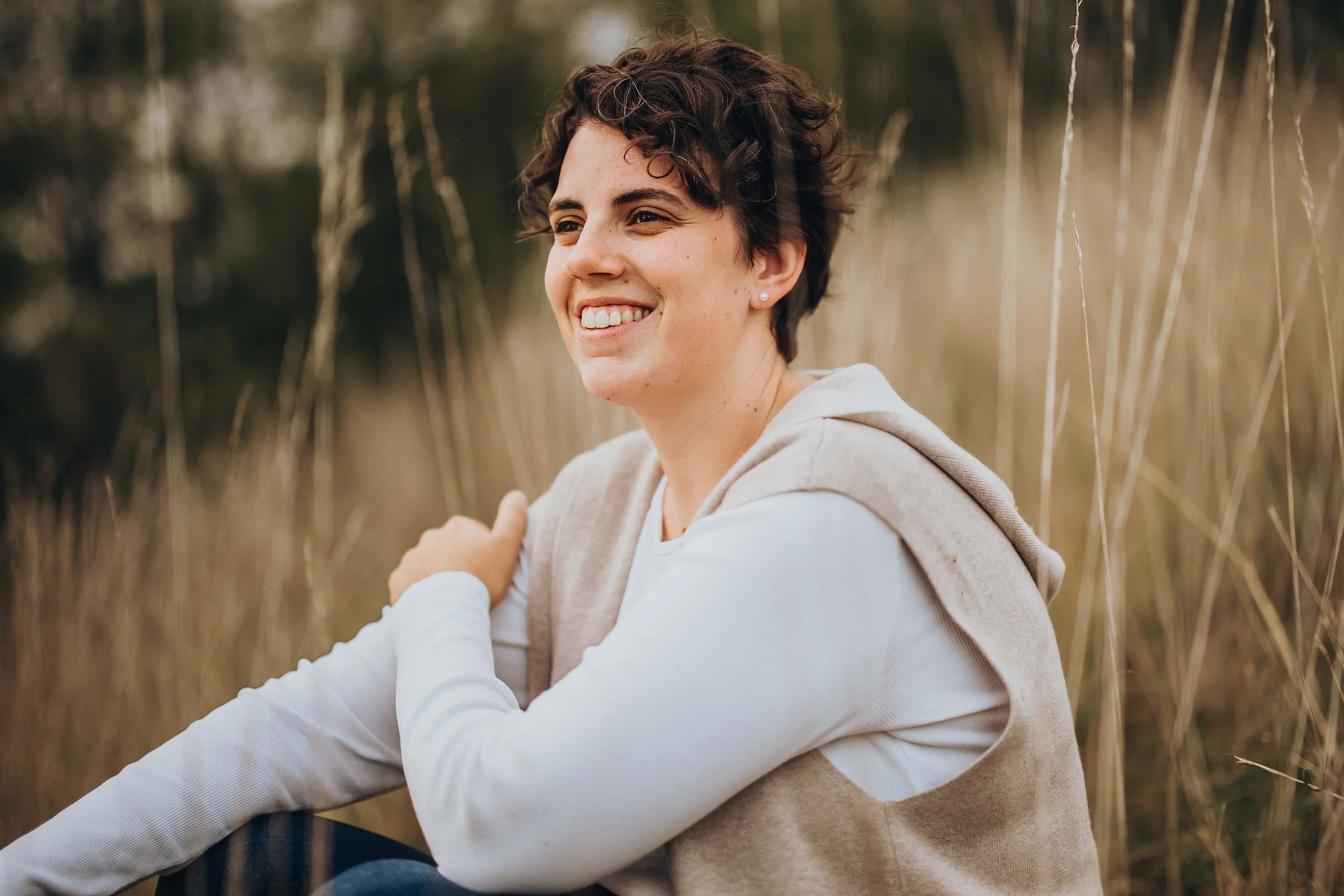 Alexandra Stagl sitzt in einer Wiese, trägt eine weiße Jacke und einen beigen Pullover, umgeben von hohen Gräsern.