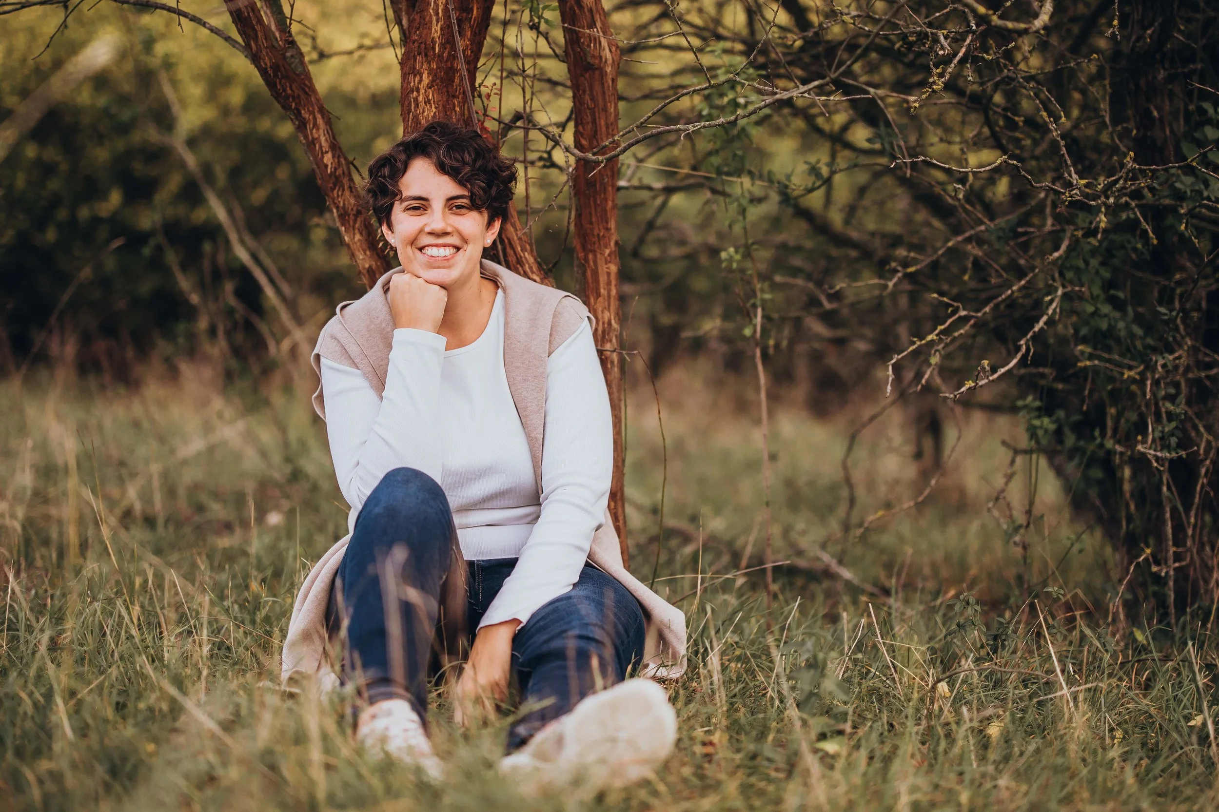 Alexandra Stagl sitzt im Gras in der Nähe eines Baumes im Freien an einem sonnigen Tag.