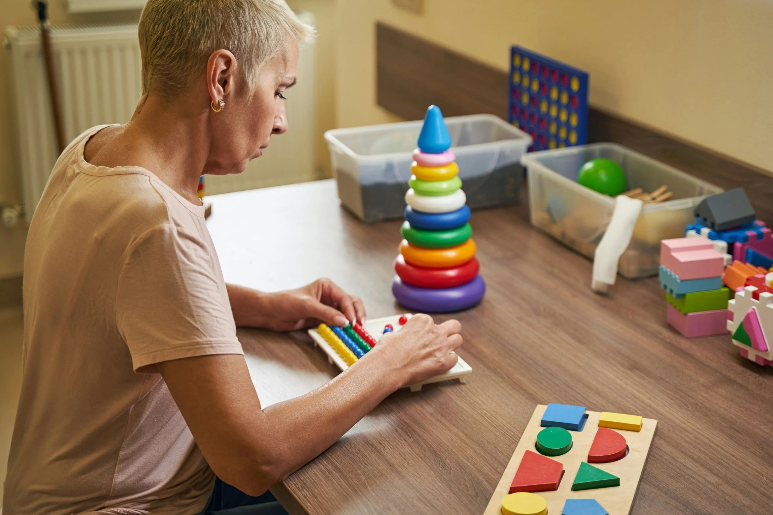 Eine ältere Frau spielt mit bunten Lernspielzeugen an einem Tisch, darunter ein aufstellbares Stapelspiel, ein Abakus und geometrische Formen miteinander.