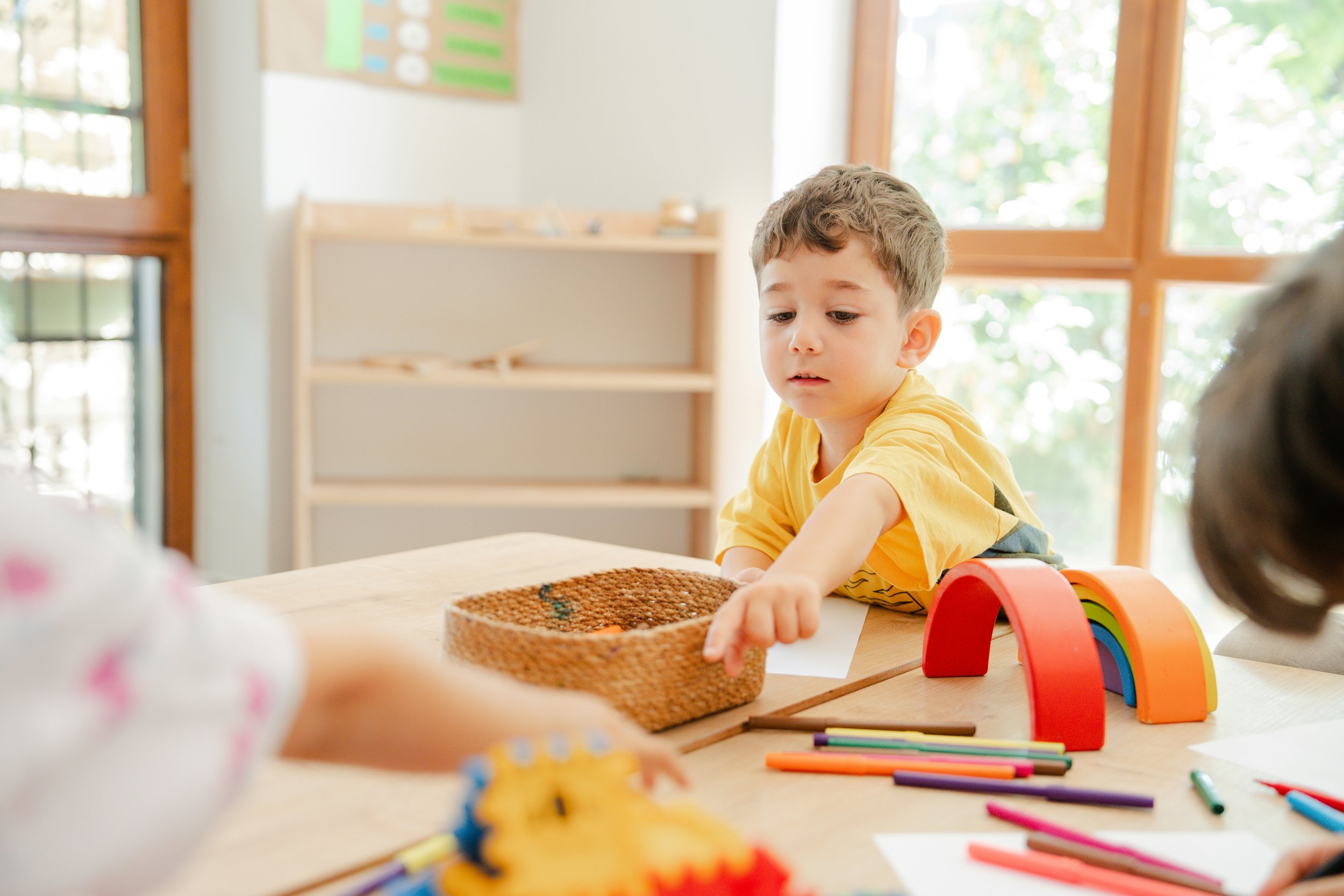 Ein Junge in einem gelben T-Shirt, der mit bunten Stiften und Spielzeug auf einem Tisch im Kindergarten spielt.