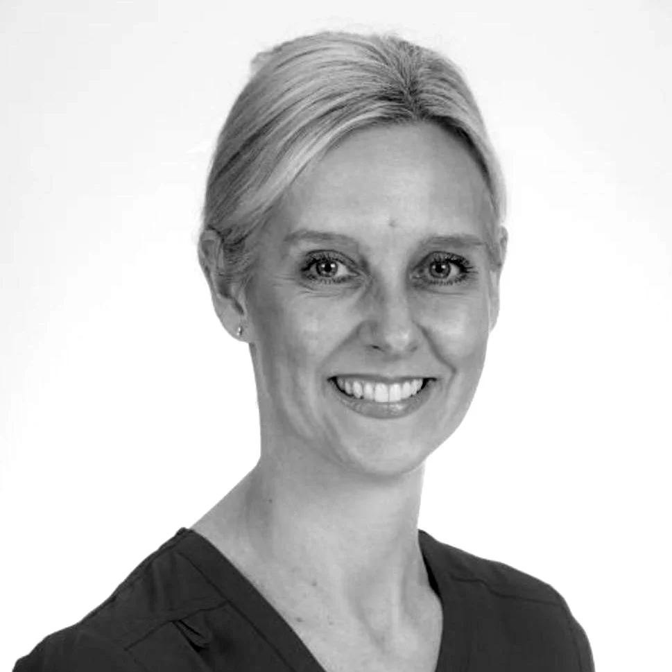 A woman with blonde hair wearing a blue medical scrub top, smiling in front of a plain white background.