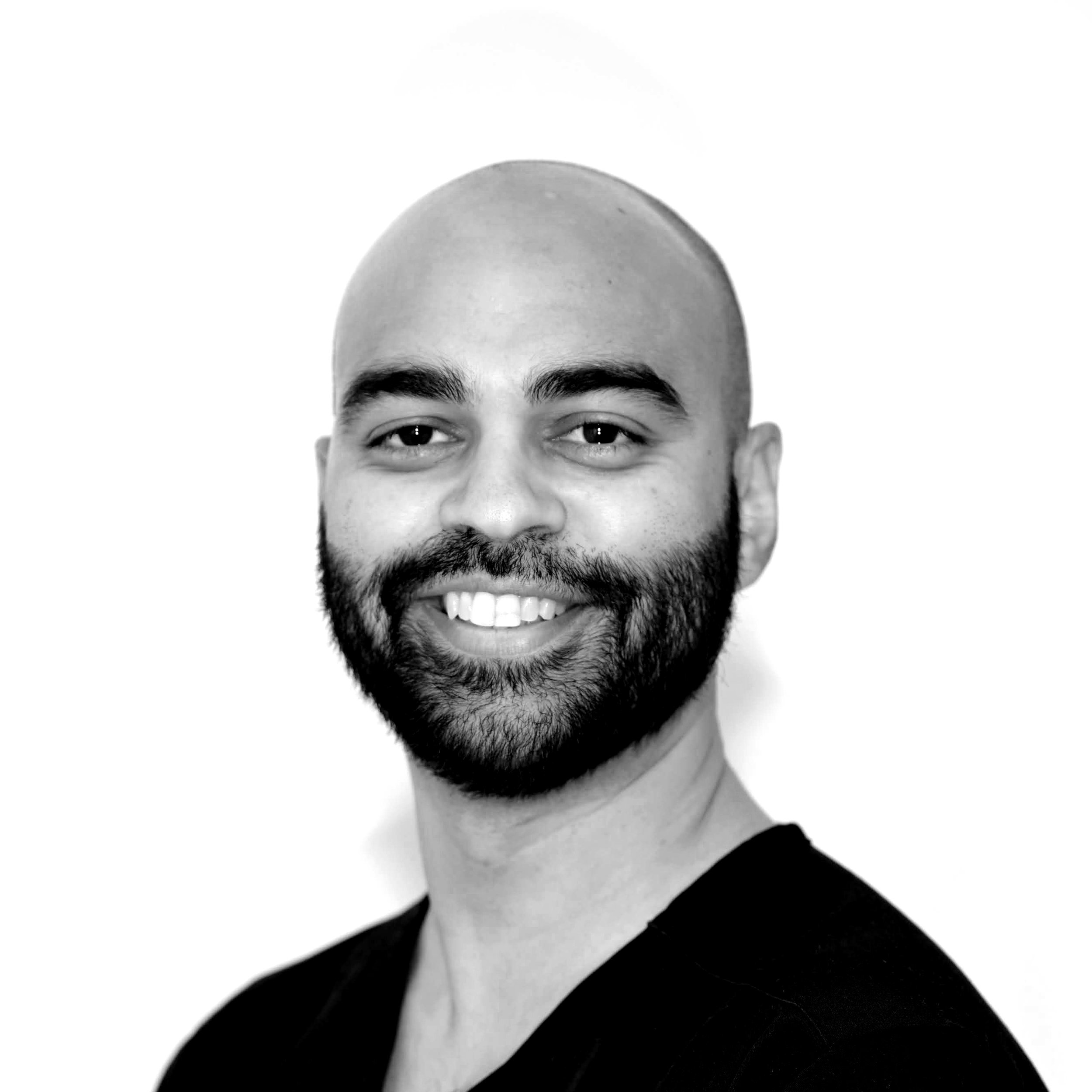 A man with glasses, a beard, and a shaved head smiling outdoors, wearing a blue shirt.