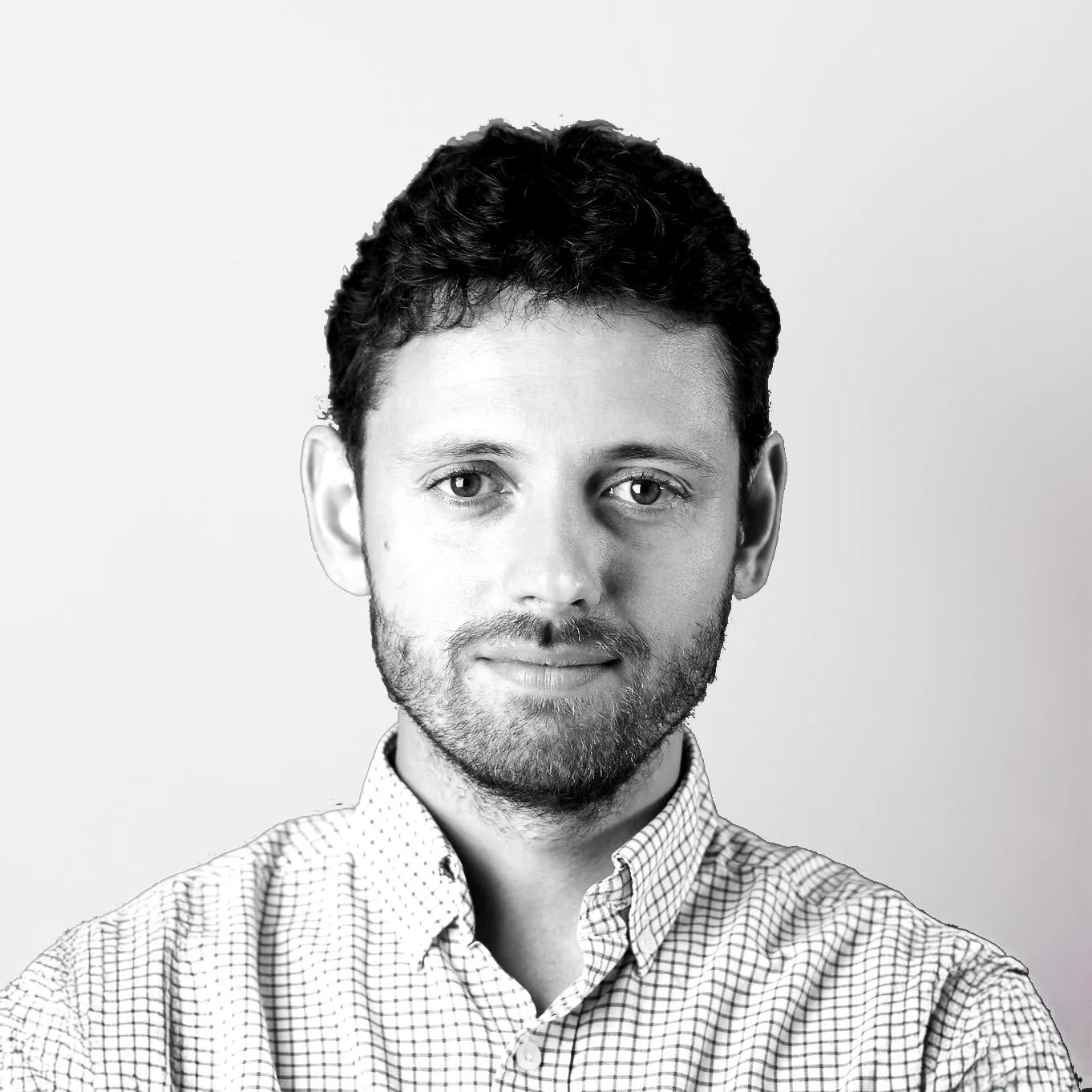 Black and white portrait of a man with short, dark, curly hair and a beard, wearing a checkered shirt, against a plain background.