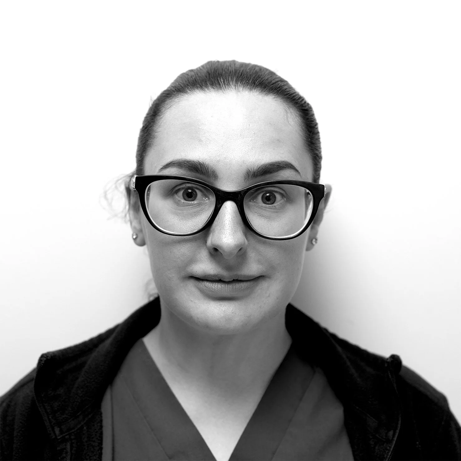 Black and white portrait of a woman with glasses, slicked-back hair, wearing scrubs and a jacket against a plain white background.