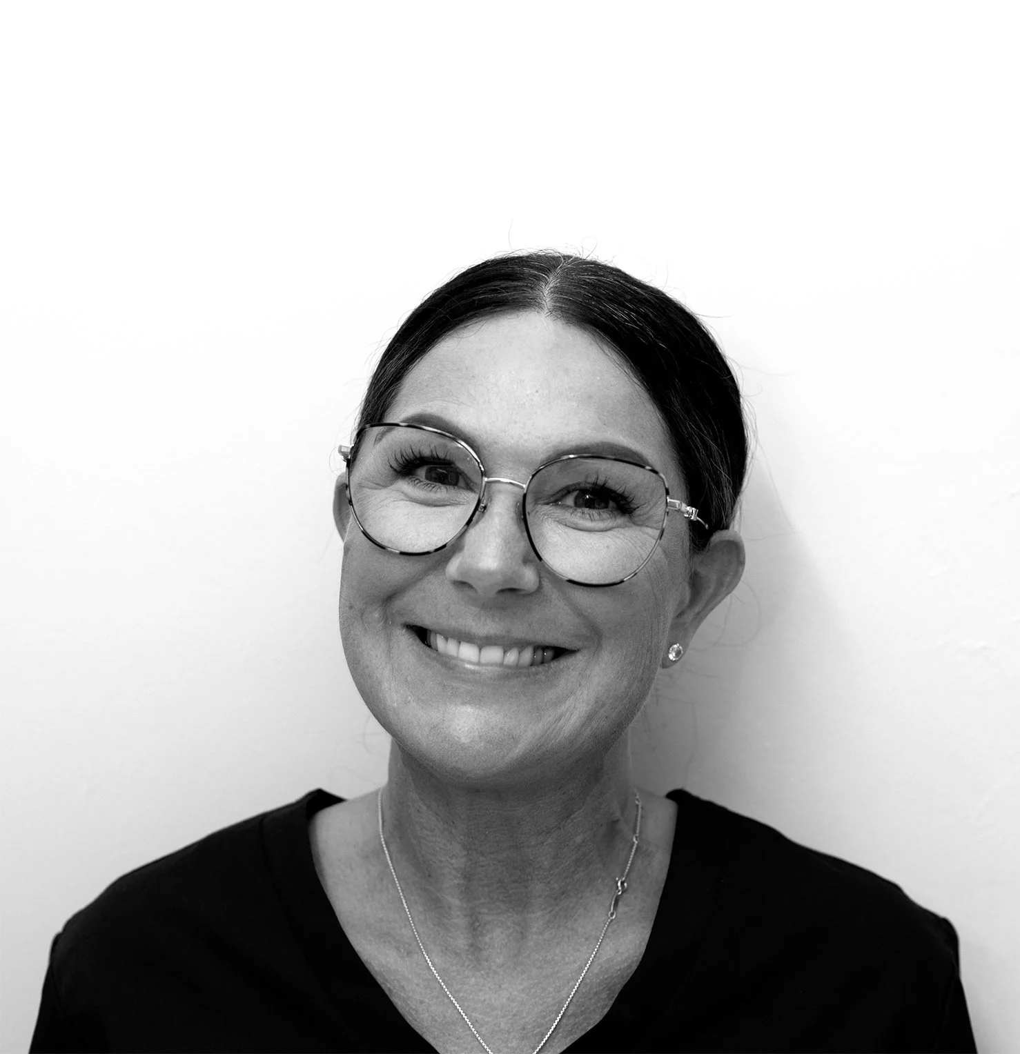 Black-and-white photo of a smiling woman with glasses, dark hair pulled back, wearing a necklace and small earrings, against a plain background.