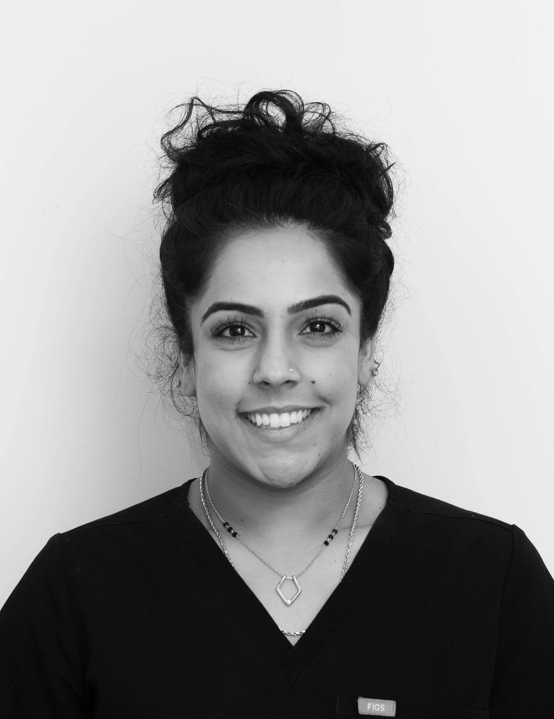 A woman with long black hair smiling at the camera, wearing a navy blue scrub top and small earrings.