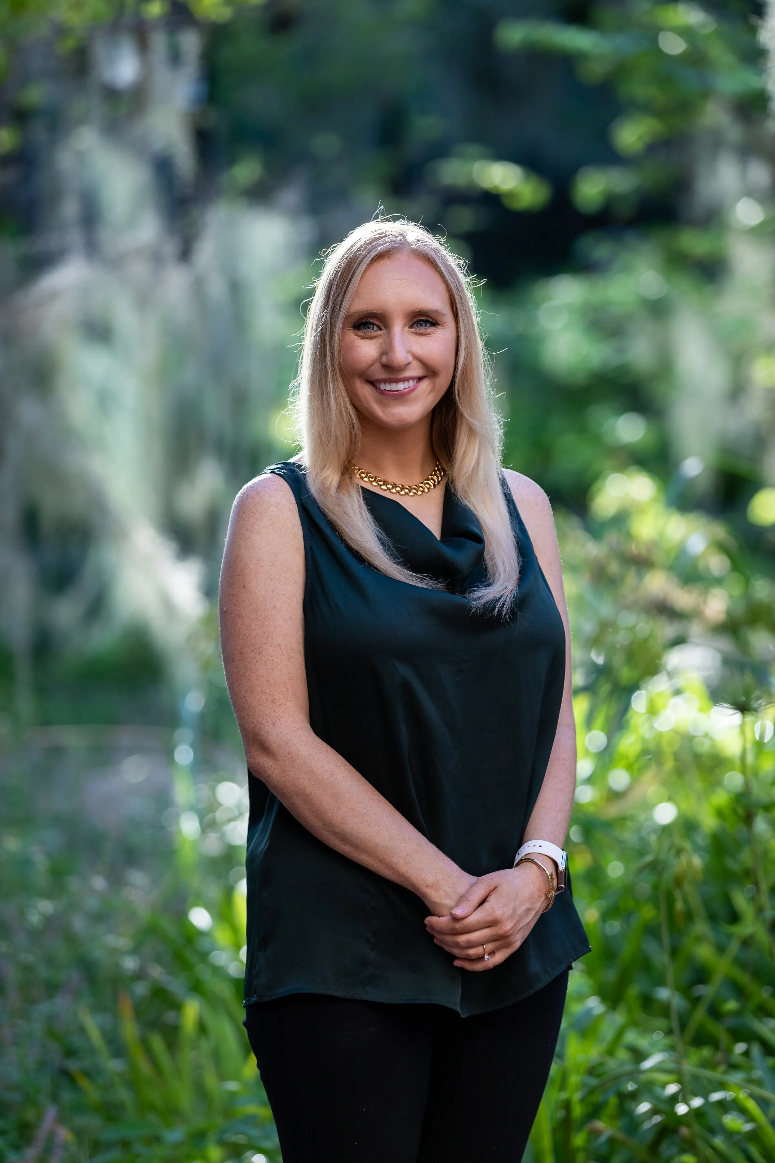 A woman with blonde hair, wearing a sleeveless black top with a gold chain necklace, standing outdoors in a lush green area with trees and plants, smiling at the camera.
