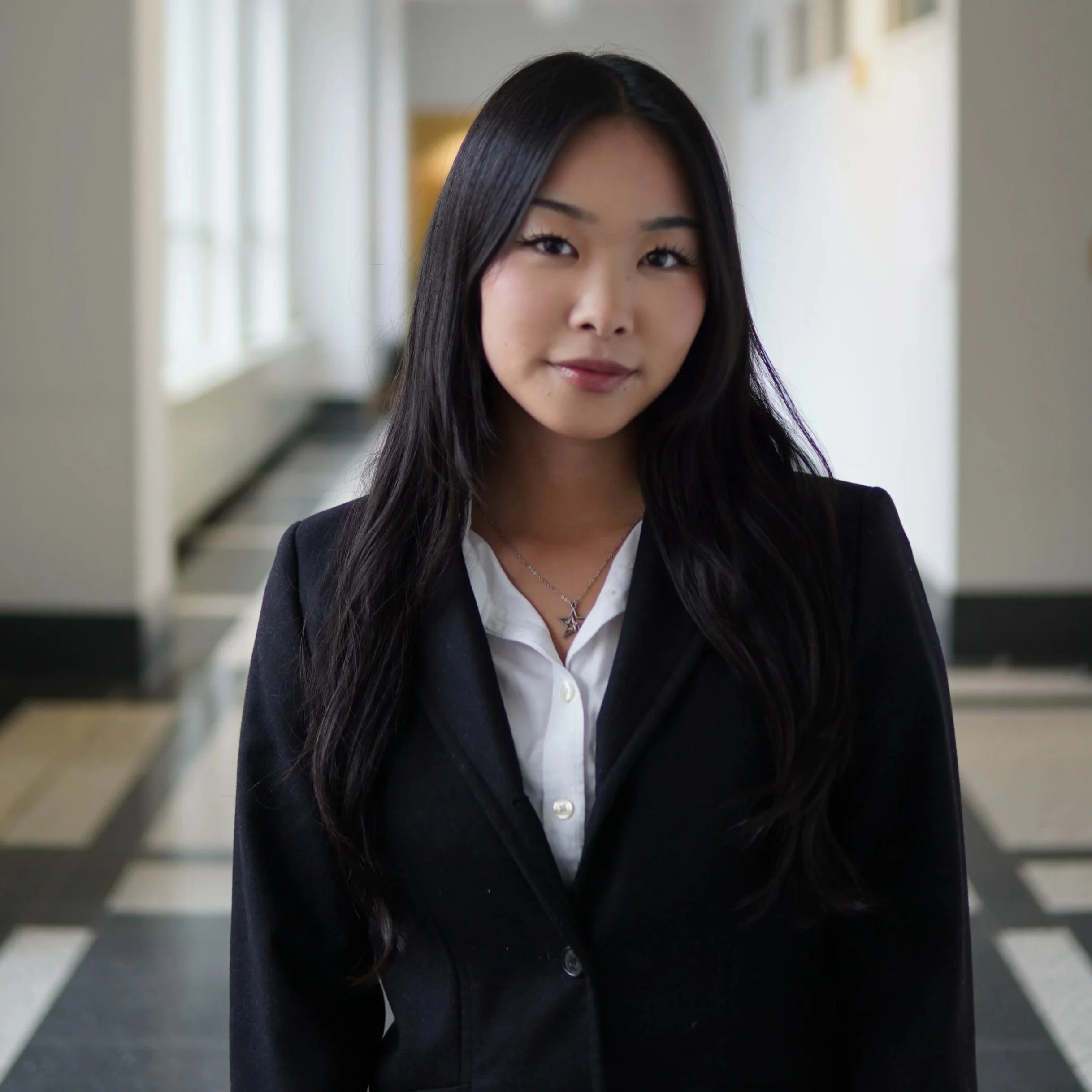A young woman with long blonde hair and blue eyes, wearing a white top and black blazer, standing in a bright, modern office corridor.