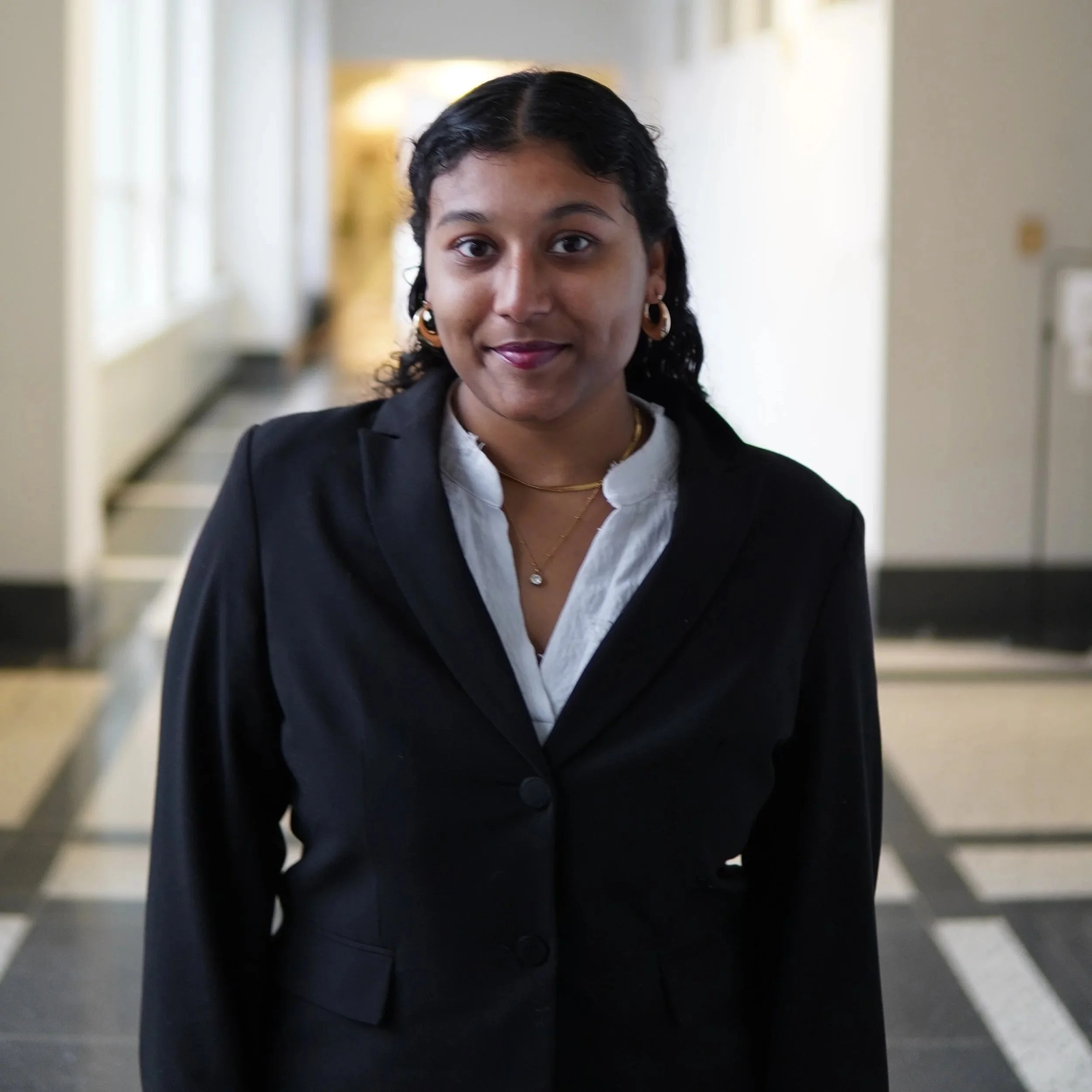 A woman with shoulder-length blonde hair, wearing glasses, a white turtleneck, and a black blazer, stands in a bright corridor with large windows and tiled floor.
