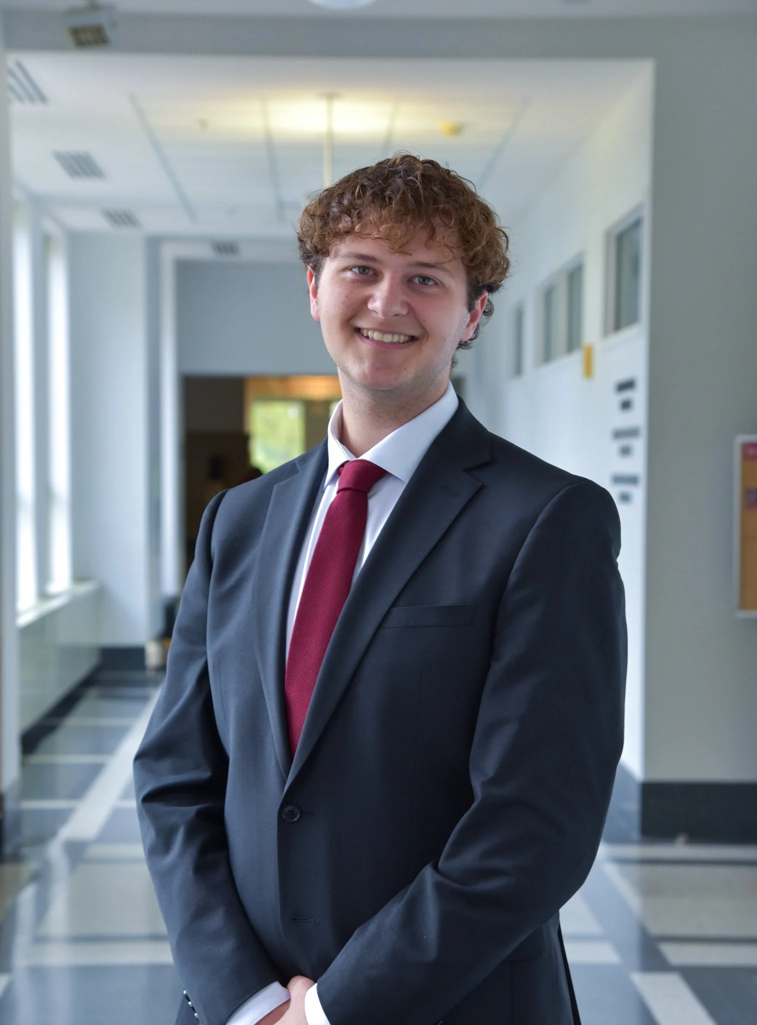 A young woman with long dark hair, wearing a light gray blazer and a black top, smiling in a brightly lit indoor corridor with large windows.