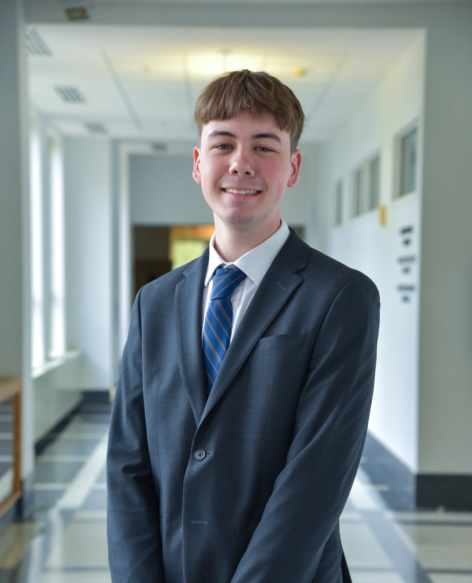 A young man in a dark suit, light blue shirt, and red tie standing in a bright office hallway with windows and white walls.