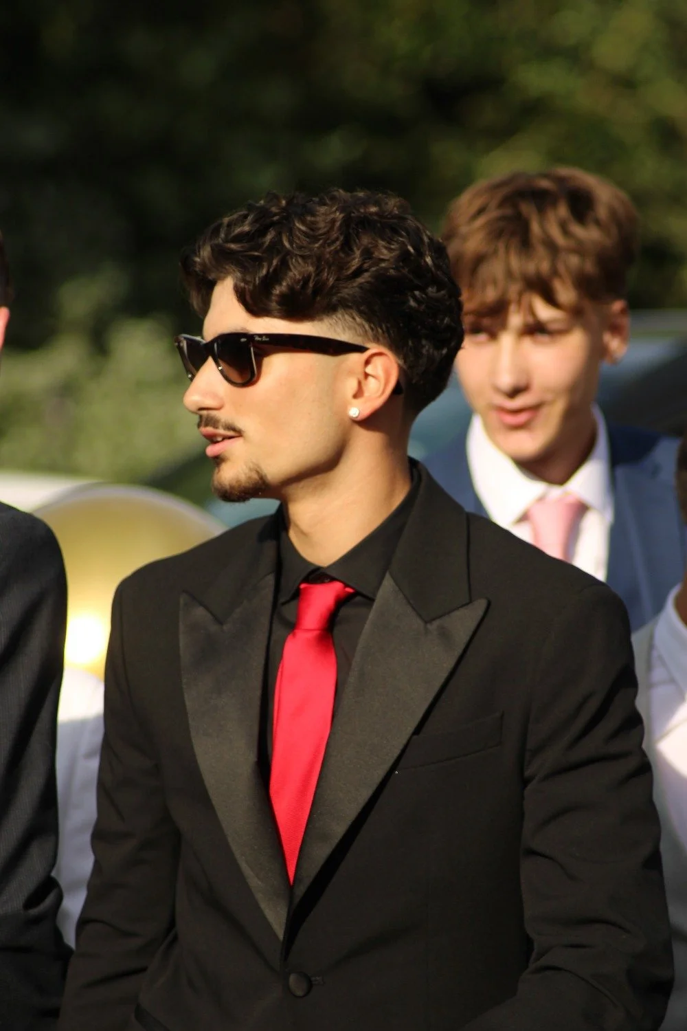 Young man in a black tuxedo with a red tie, black sunglasses, and earrings, standing outdoors with others in formal attire in the background. Prom in Pyrford, Woking, Surrey. Photographer: Blake Charles.