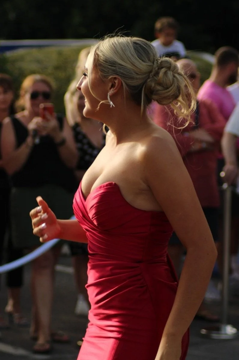 A woman in a strapless red dress with her hair styled in an elegant updo, smiling and surrounded by people at an outdoor event. Prom in Pyrford, Woking, Surrey. Photographer: Blake Charles.