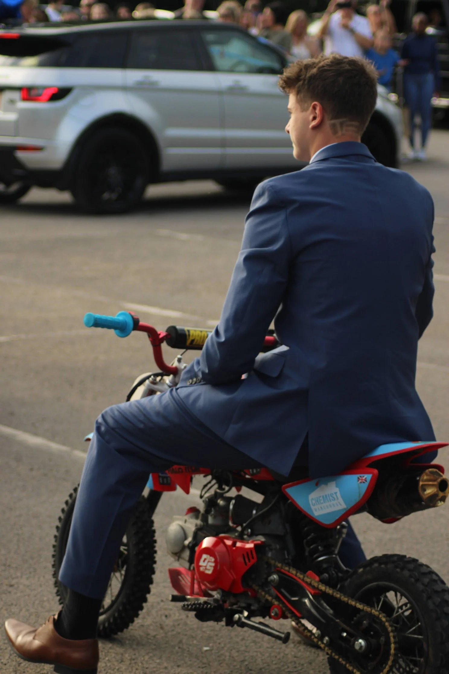 A young man at a Prom in Pyrford, Woking, in a blue suit sitting on a small red dirt bike in a parking lot, with a crowd and a silver SUV in the background. Photographer: Blake Charles