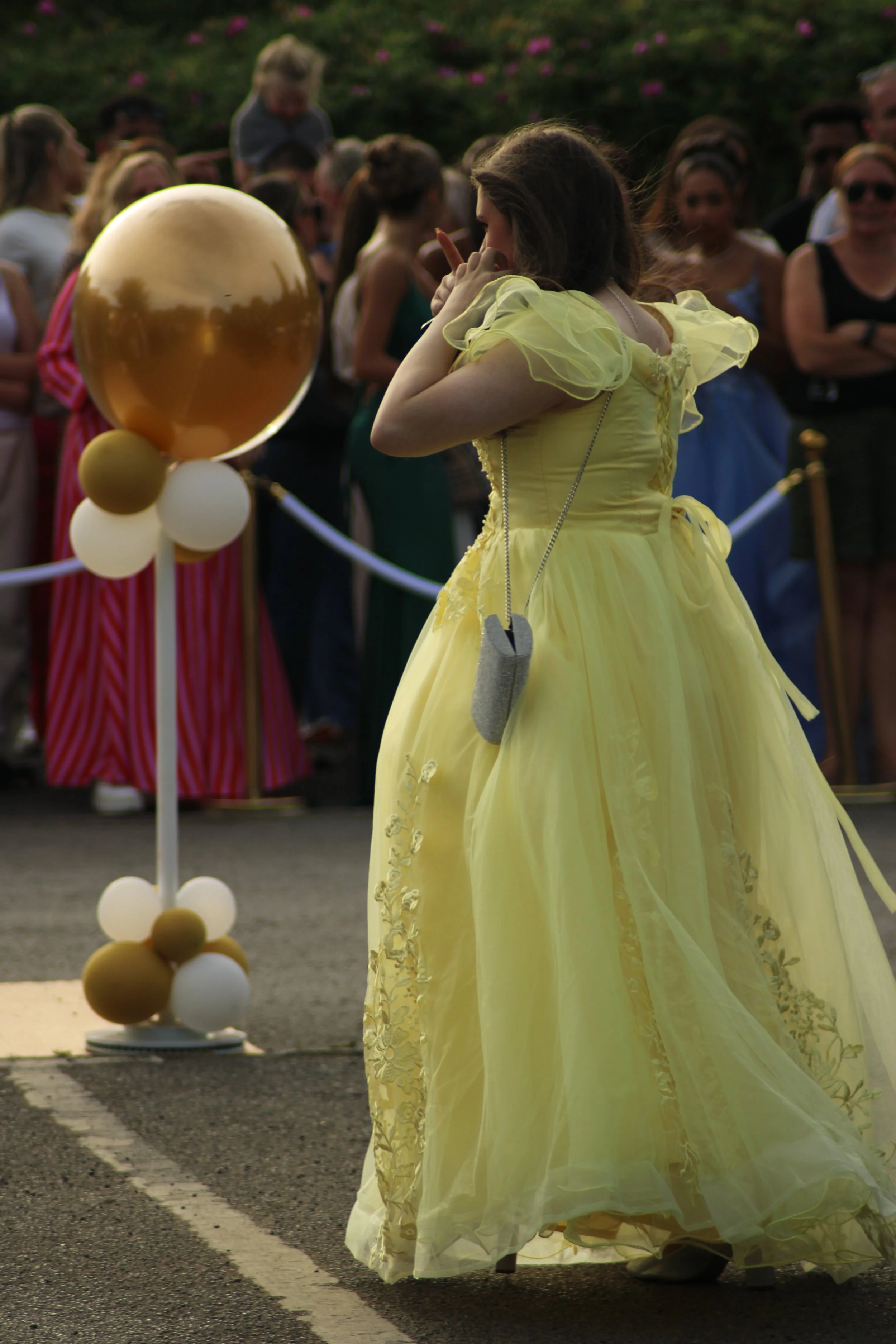 A young woman in a yellow gown standing on the street at a high school prom in Pyrford, Woking, surrounded by a crowd of onlookers, with balloons nearby. Photographer: Blake Charles.