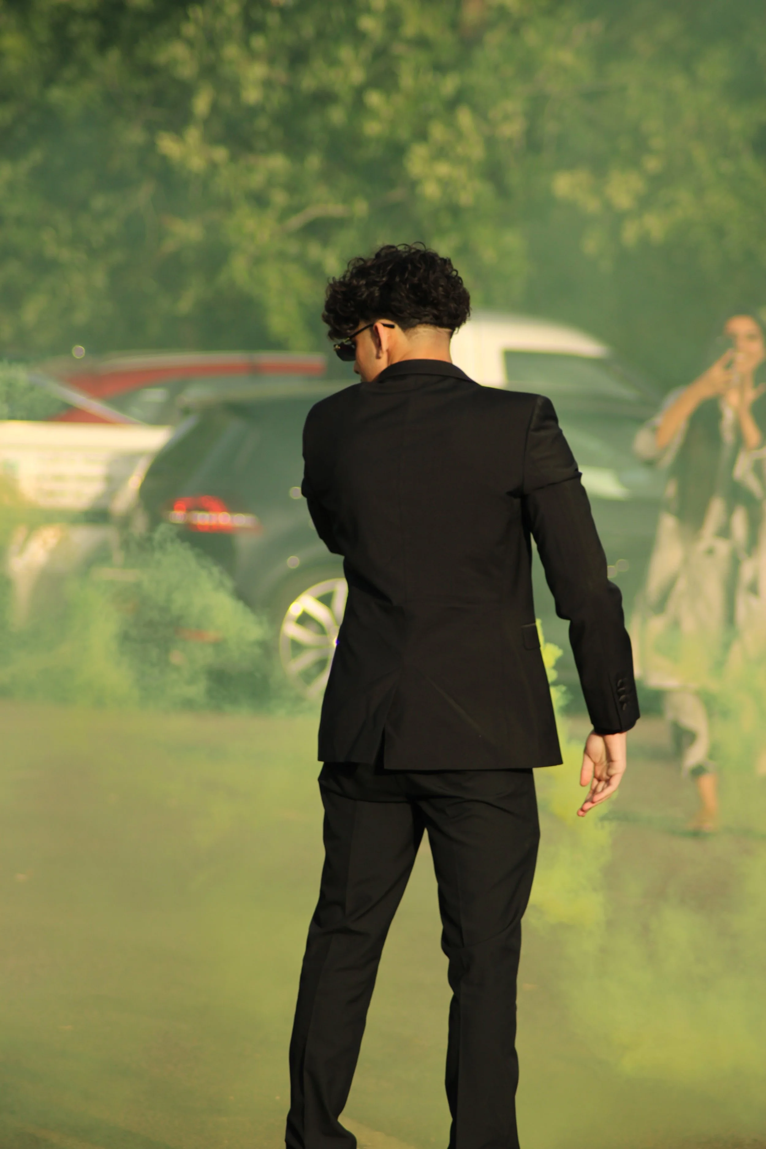 A man in a black suit with curly dark hair standing outdoors, with cars and trees in the background, and a woman taking photos to the right. Prom in Pyrford, Woking, Surrey. Photographer: Blake Charles.