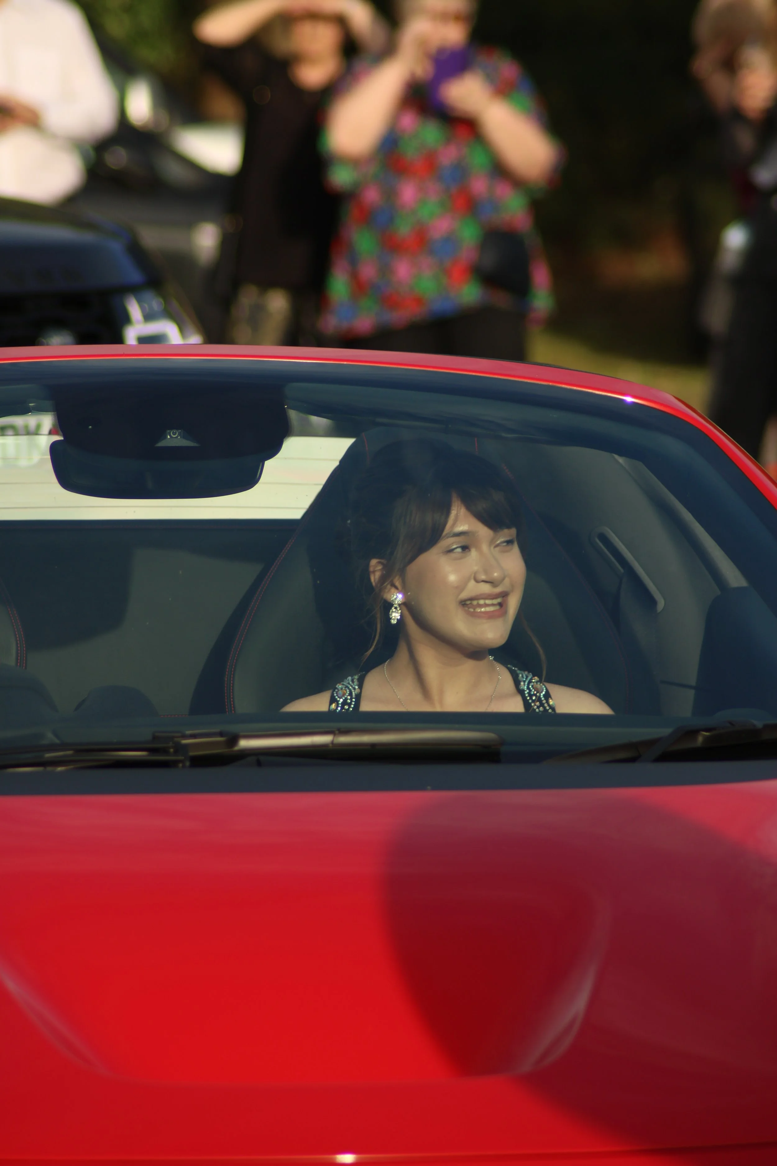 A woman sitting inside a red sports car at a Prom in Pyrford, Woking, smiling and looking out the windshield. Several people are standing outside in the blurry background. Photographer: Blake Charles