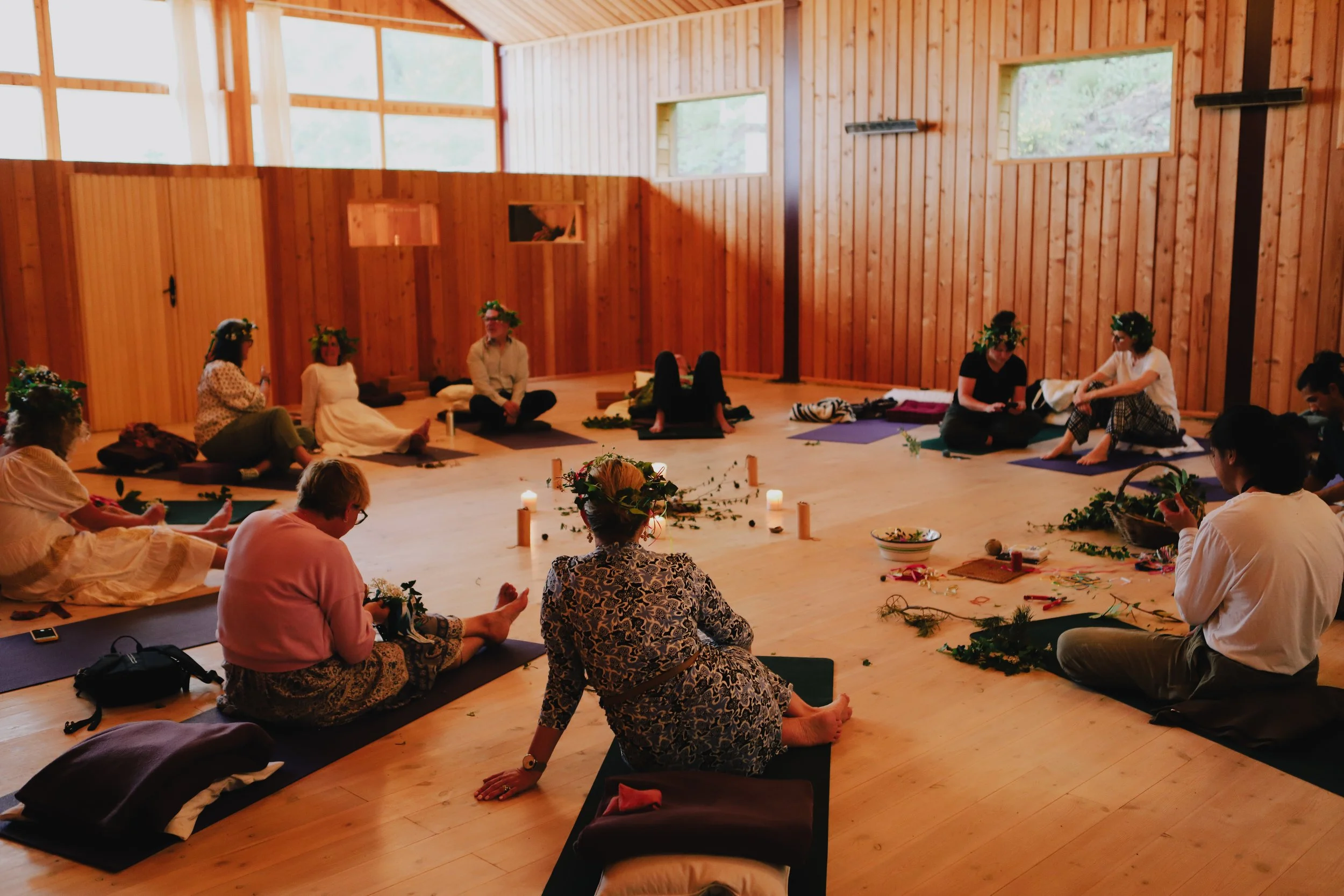 Group of people participating in a ceremony or meditation in a wooden-finished room, sitting on yoga mats and wearing flower crowns, with candles and greenery arranged on the floor.