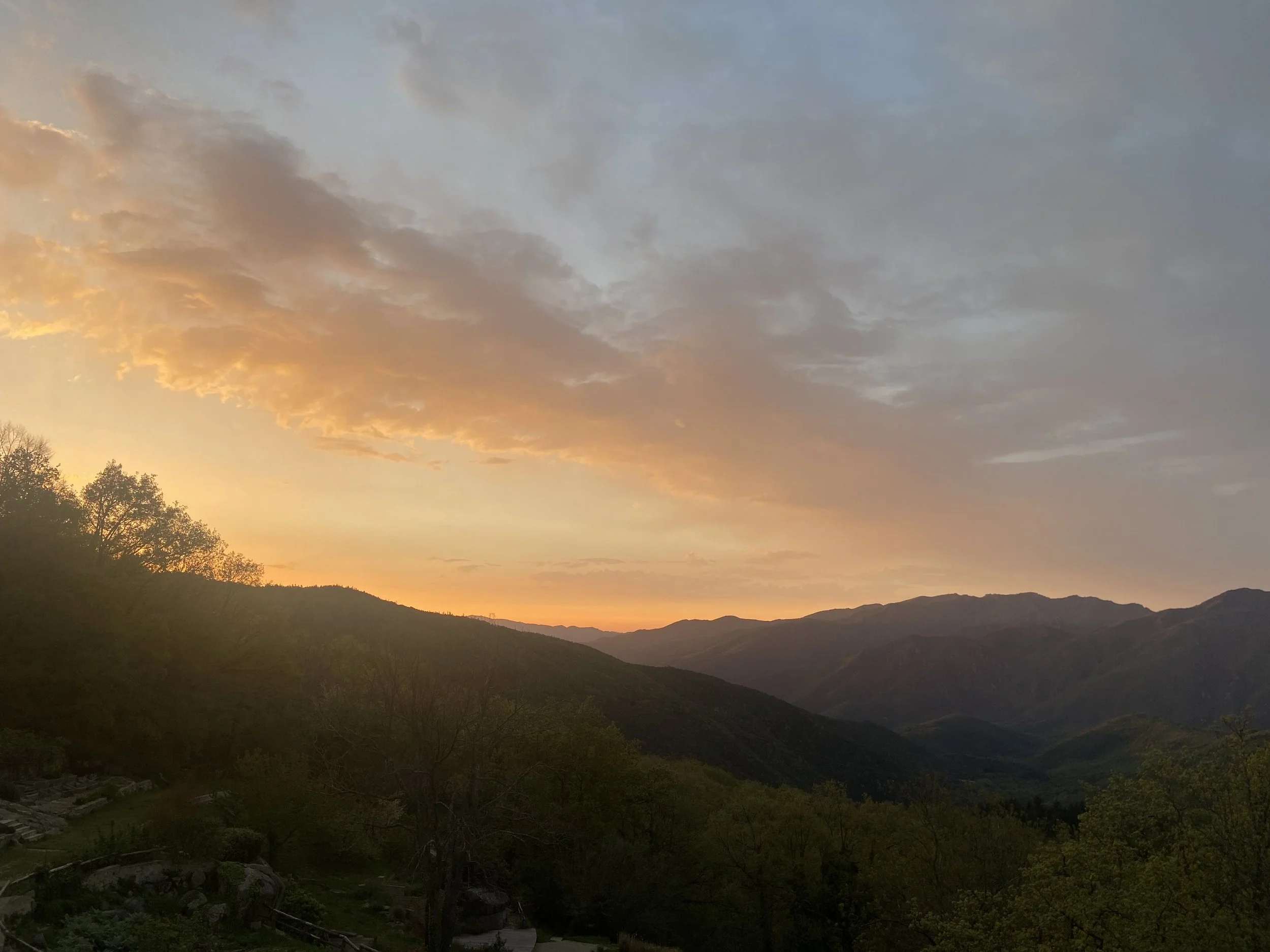 Sunset over rolling mountains with partly cloudy sky and trees in foreground.