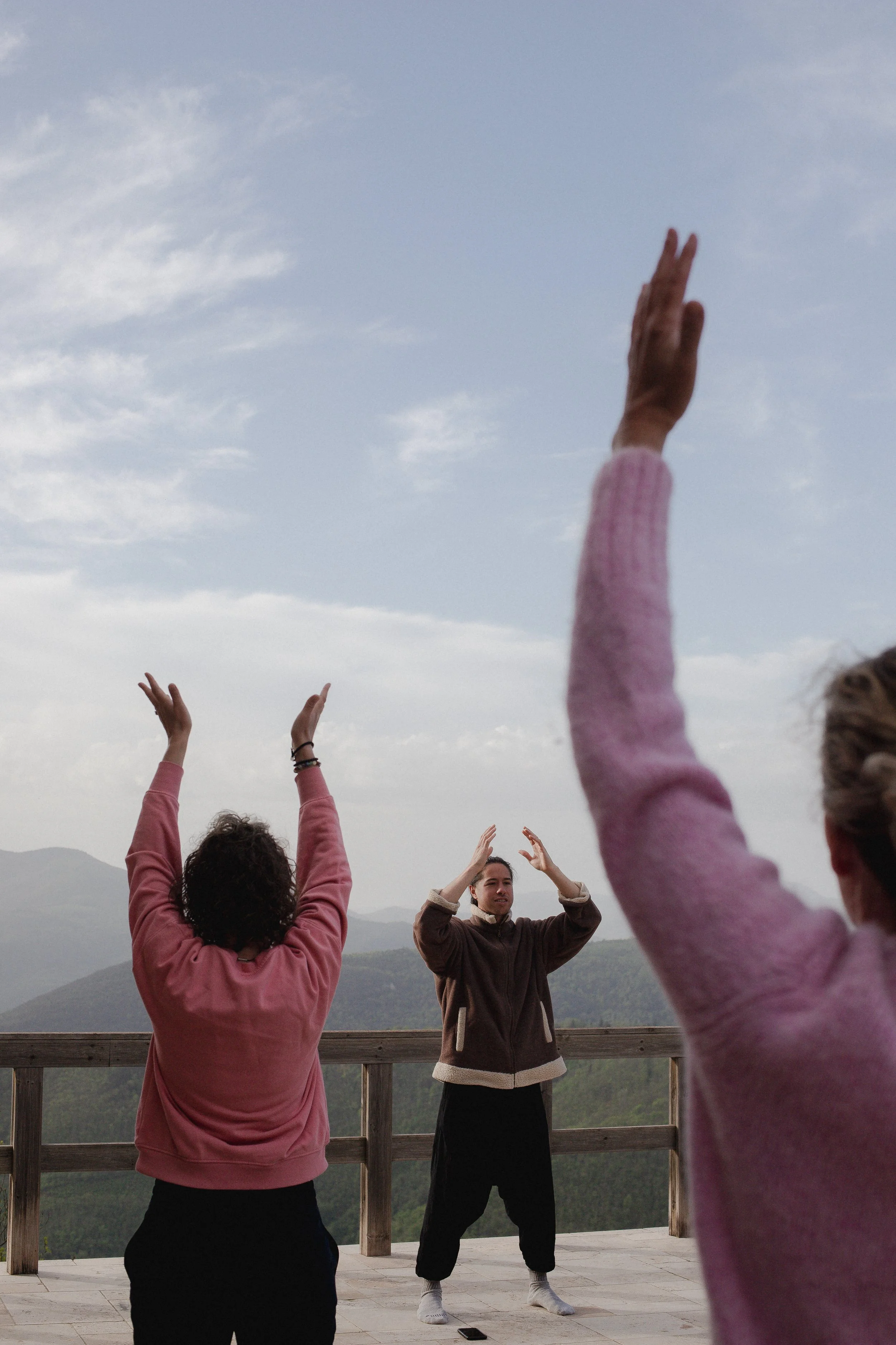 Three people practicing yoga outdoors on a wooden deck with mountain scenery, raising their arms in a yoga pose.