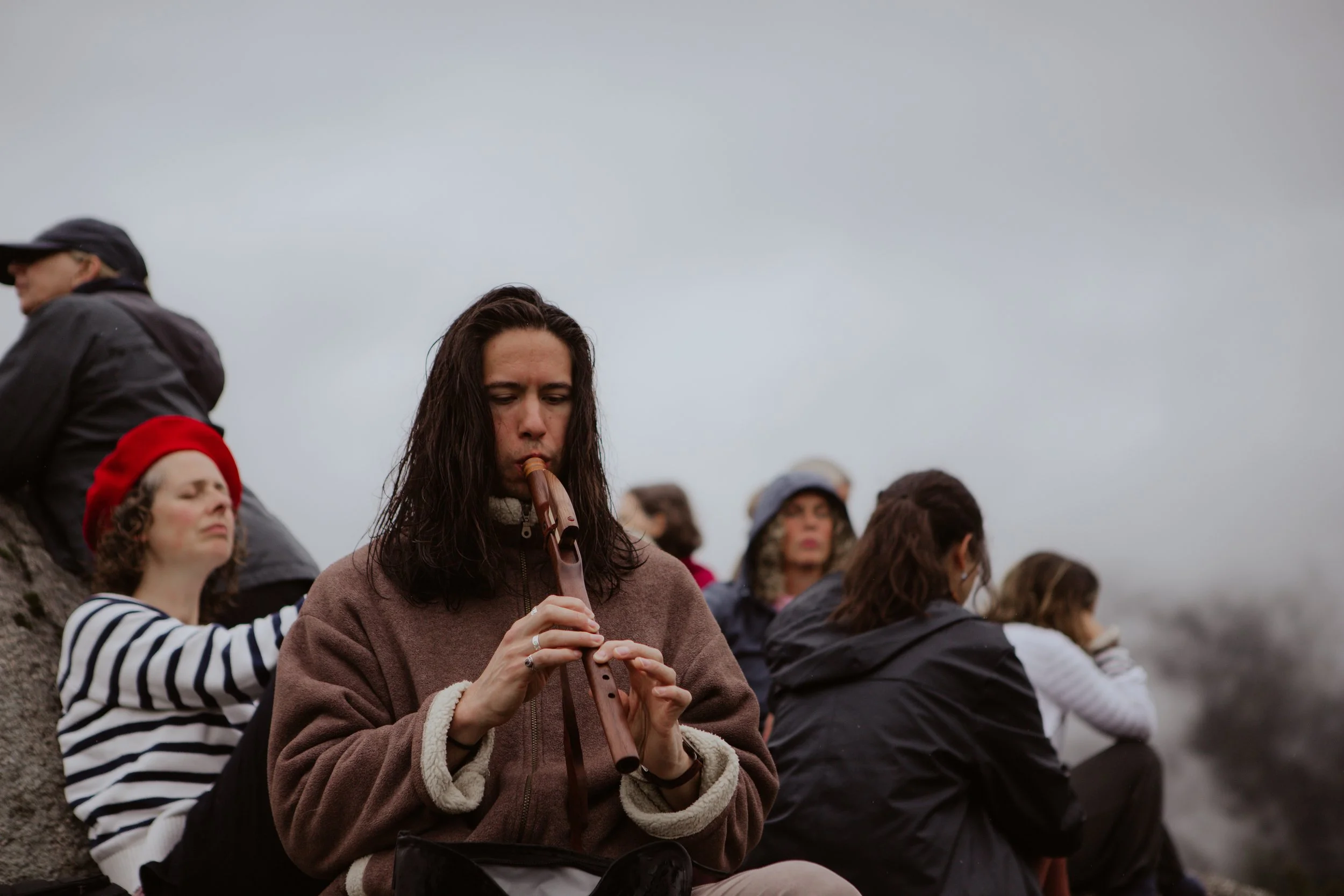 A woman with long dark hair playing a wooden flute outdoors on a cloudy day, surrounded by other people sitting and resting.