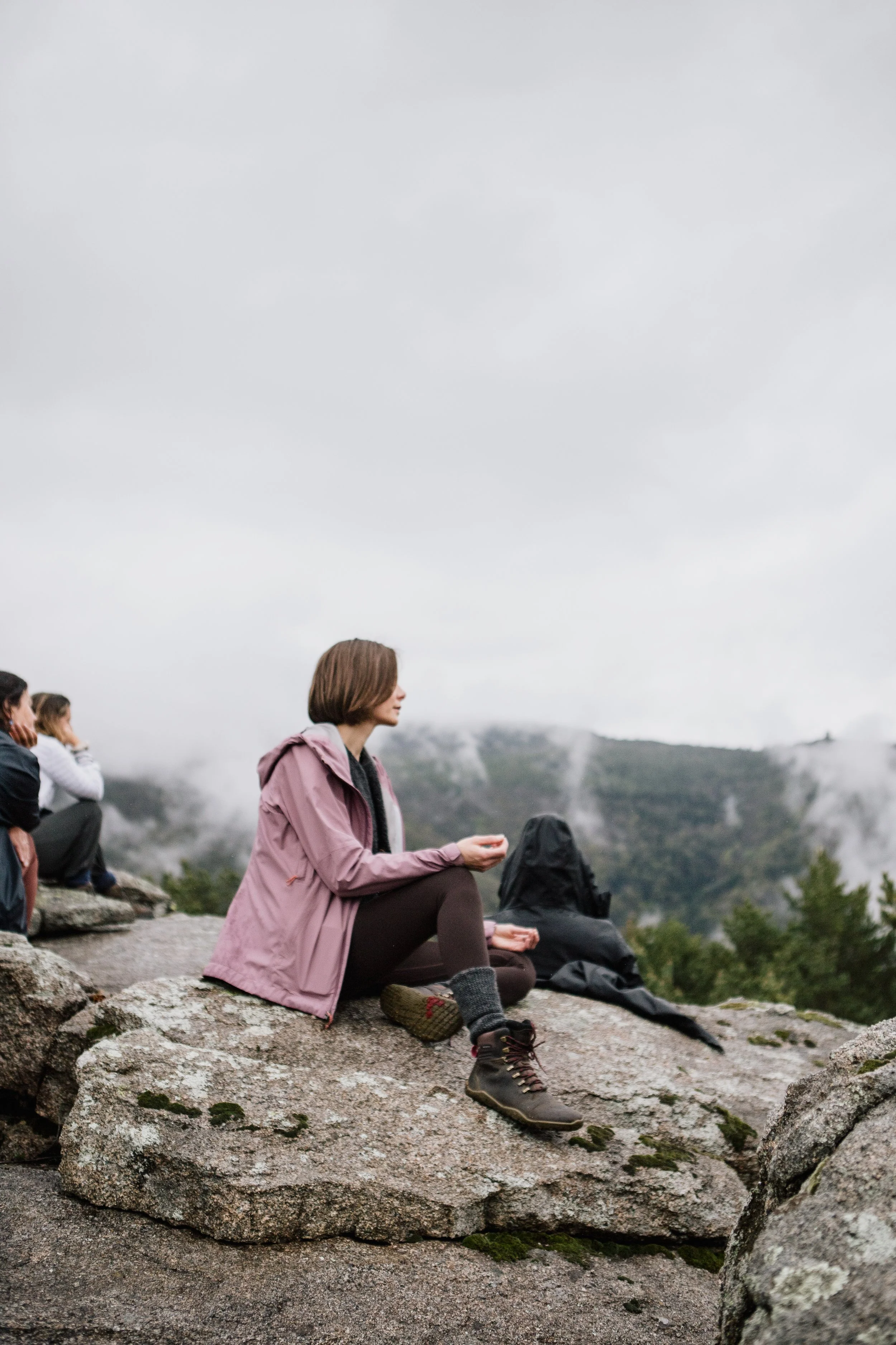 People sitting on rocks on a mountain, surrounded by fog and greenery, engaging in meditation or mindfulness practice.