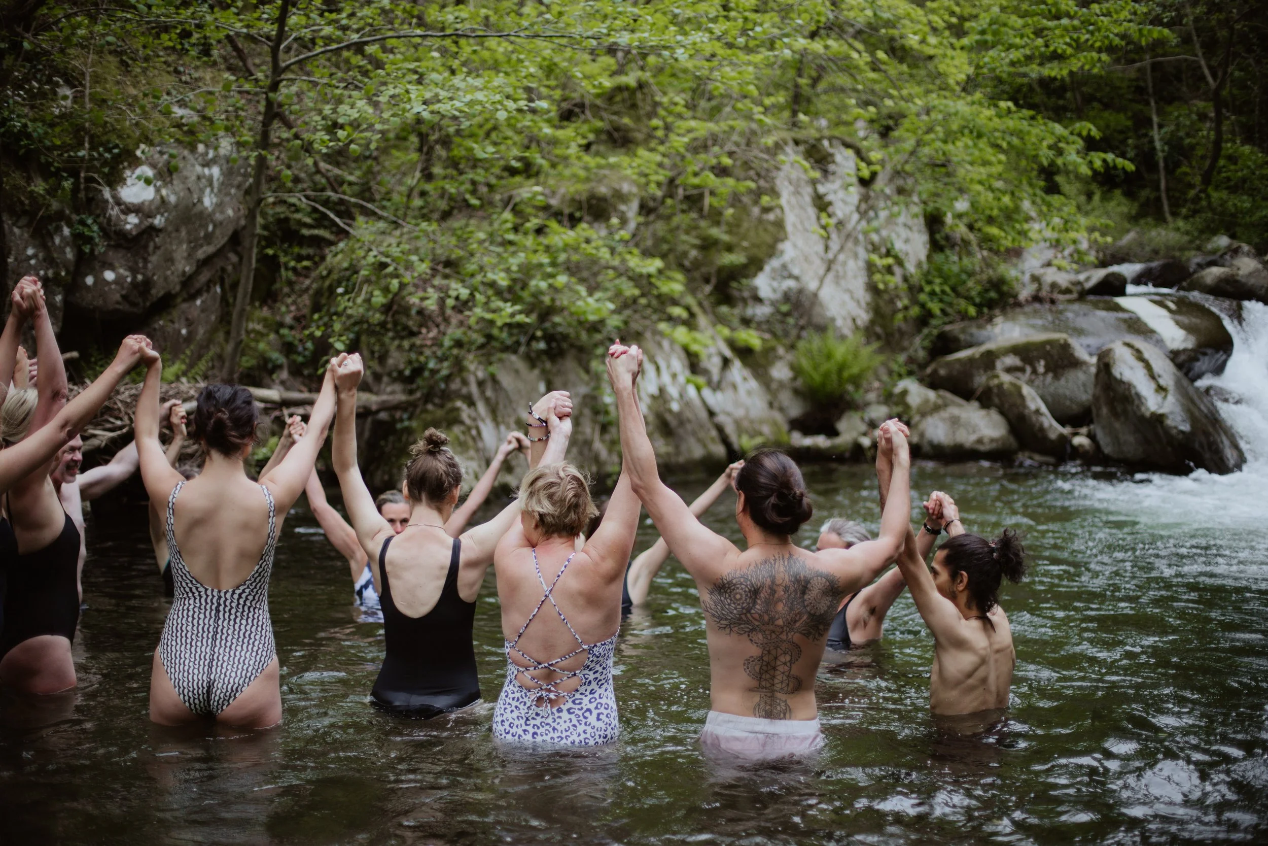 Group of people holding hands in a circle and raising their arms in a river surrounded by trees and rocks.