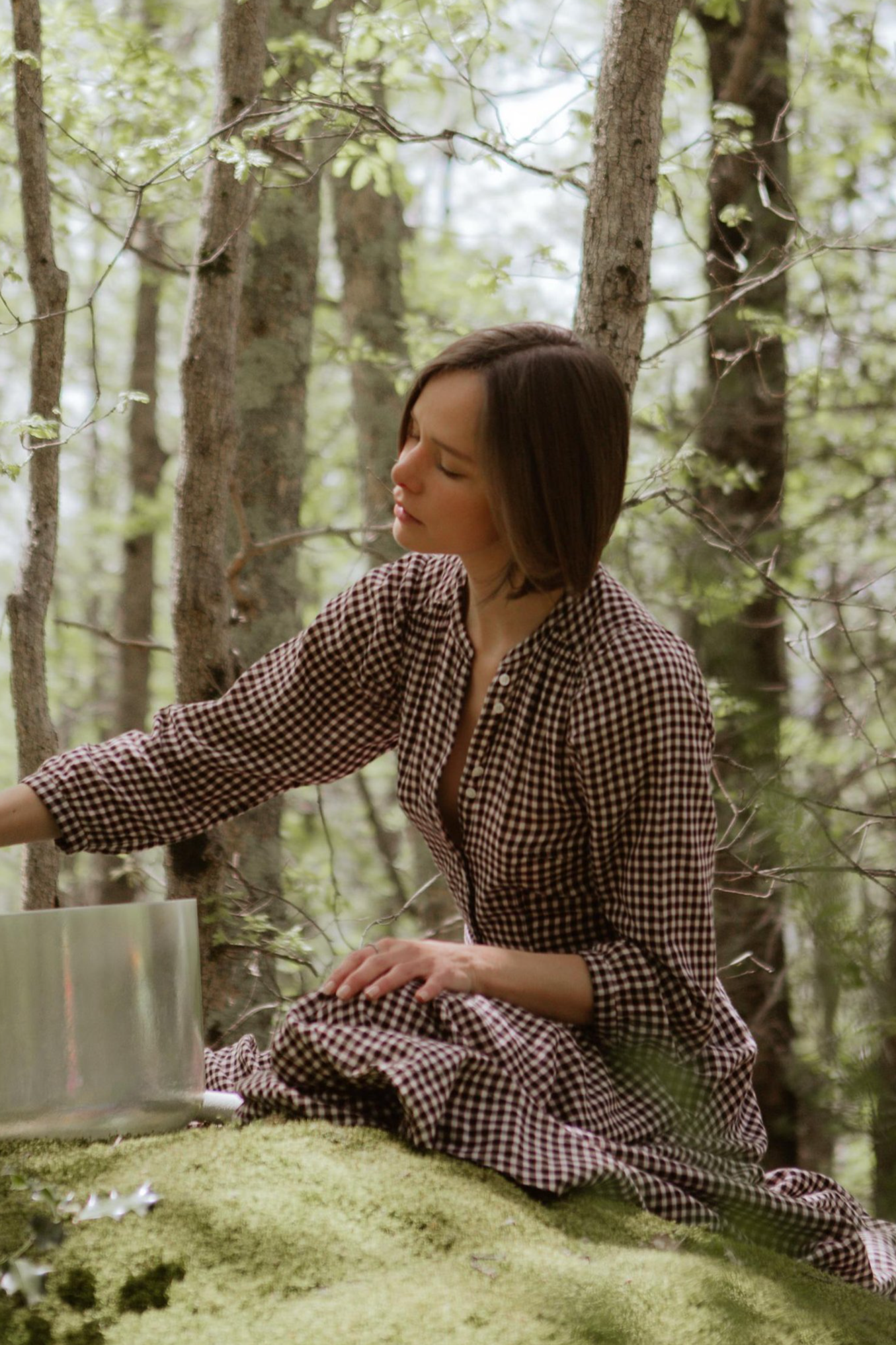 A woman with short brown hair sitting on a moss-covered surface in a forest, wearing a gingham dress, with her eyes closed and one arm extended.