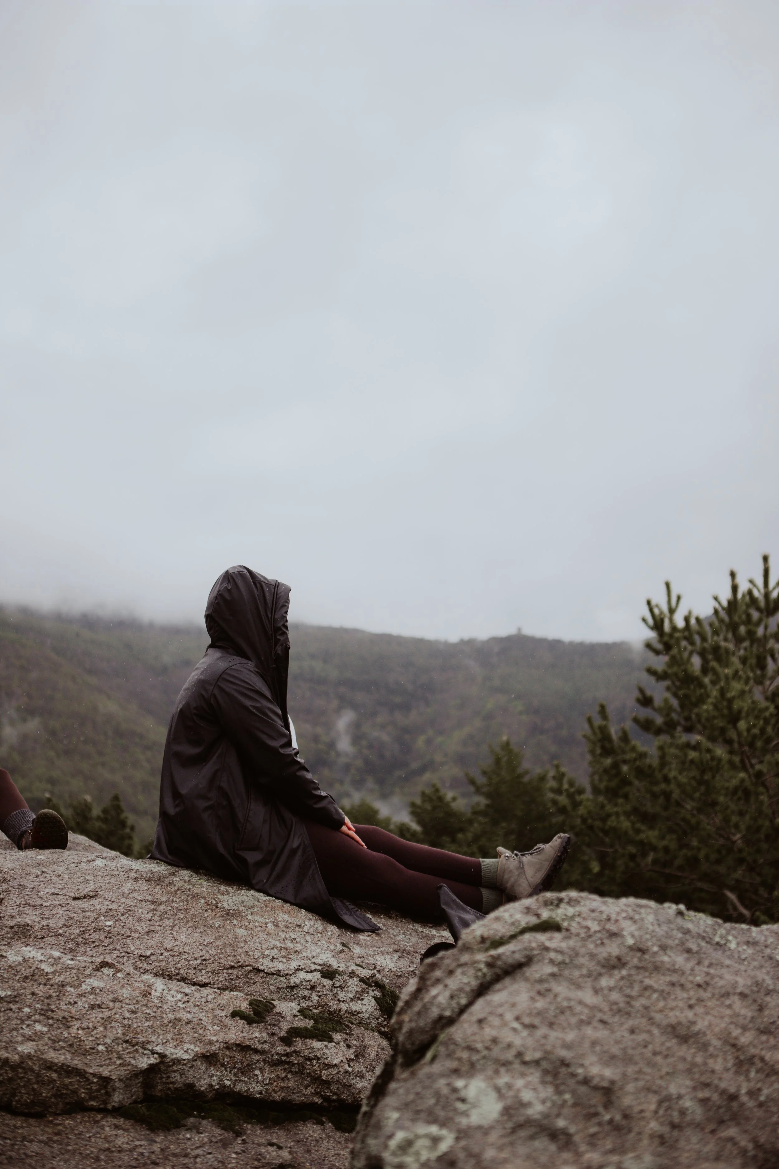 Person sitting on a large rock in a mountainous area on a cloudy day, wearing a black rain jacket with the hood up, dark pants, and boots.