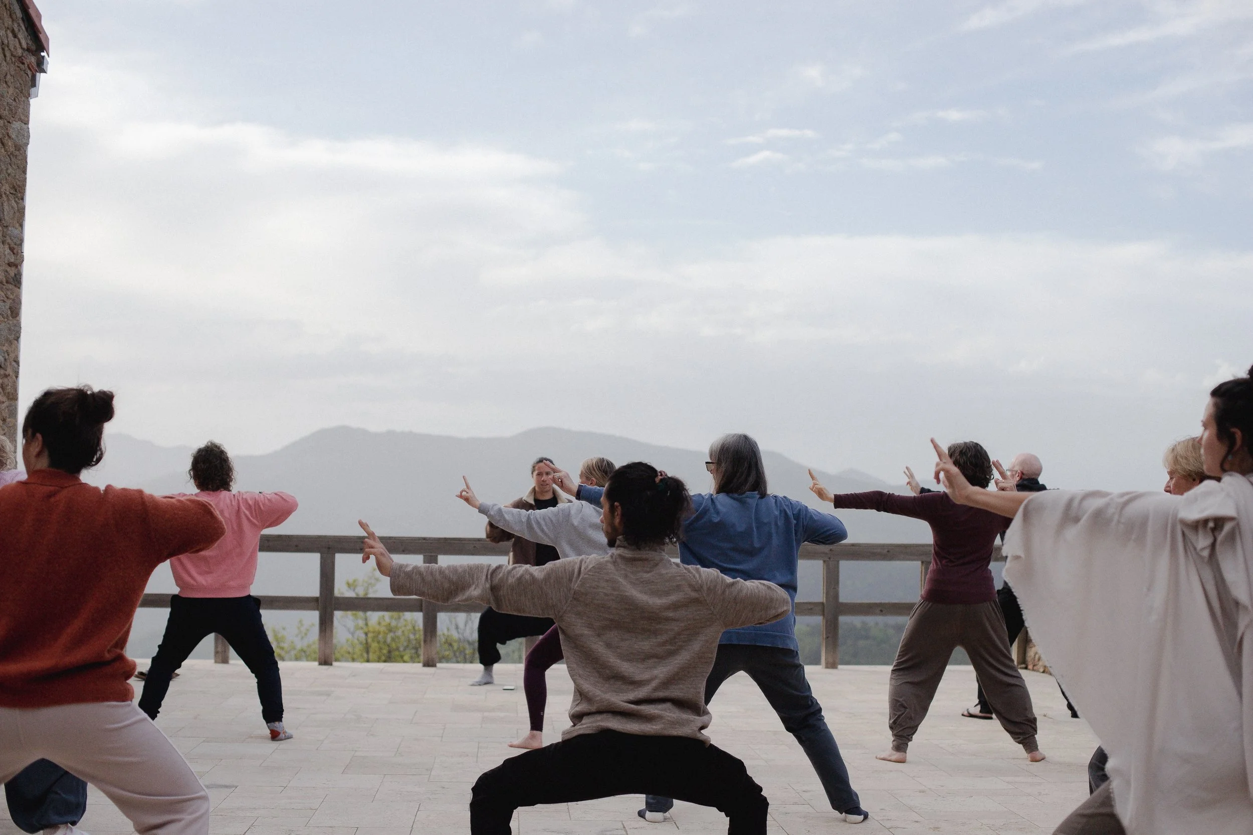 Group of people practicing yoga outdoors on a wooden deck with mountain view in the background.