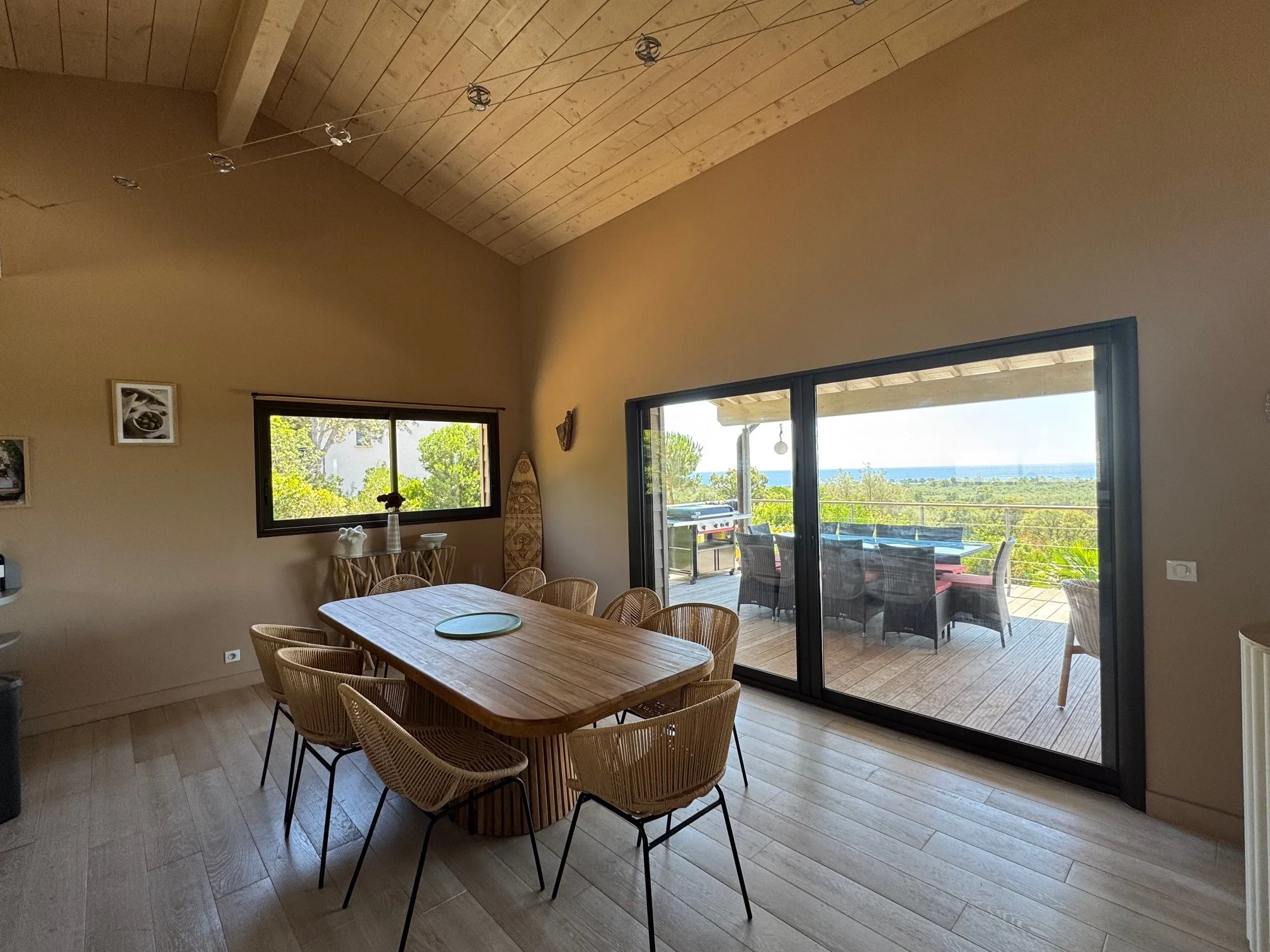 Salle à manger avec table en bois 10 pers et chaises en rotin, grande baie vitrée donnant sur une terrasse avec mobilier extérieur, vue sur la nature et la mer.