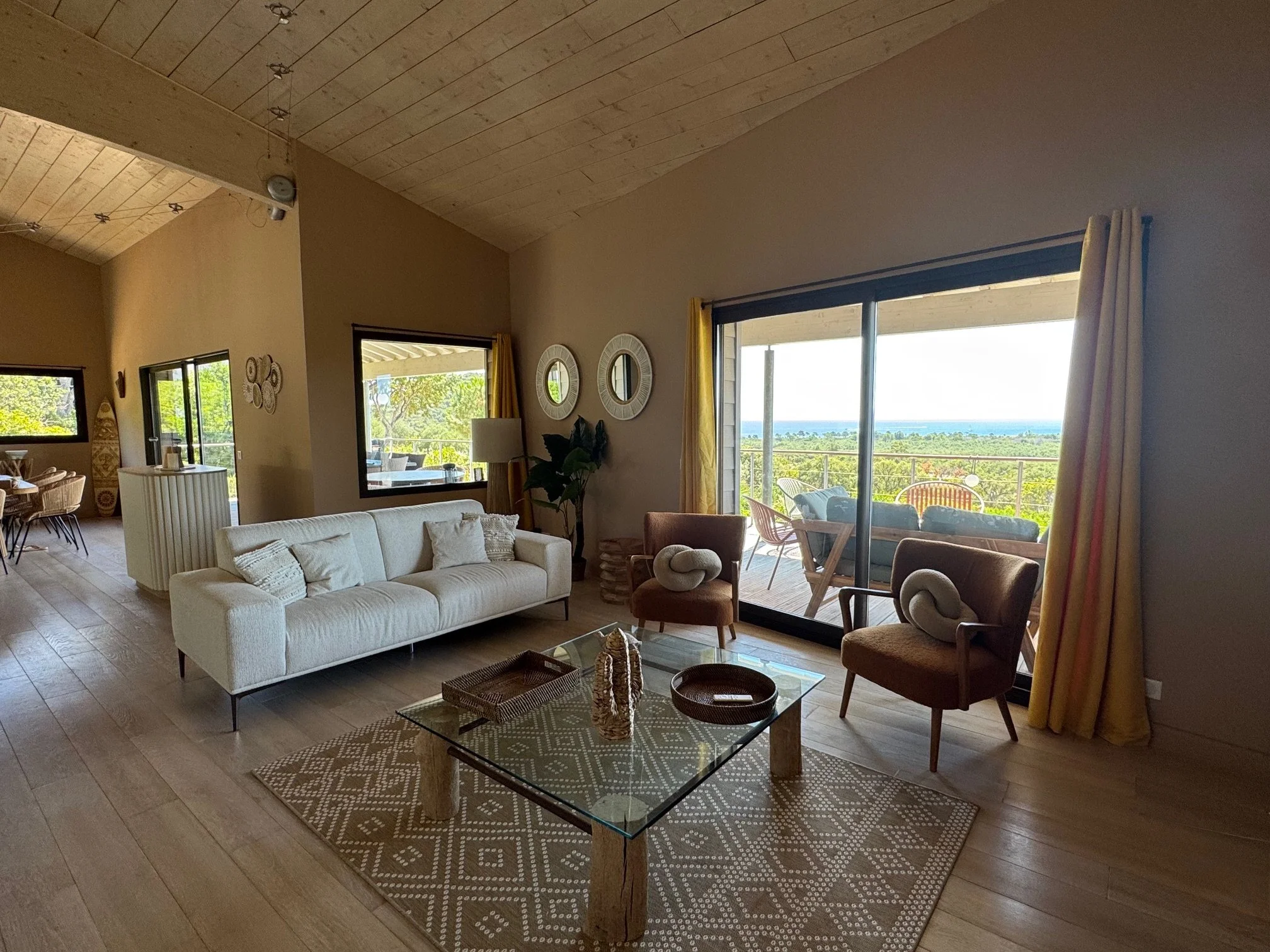 Salon moderne avec canapé beige, deux fauteuils, table en verre, vue sur une terrasse avec mobilier d'extérieur et vue mer.