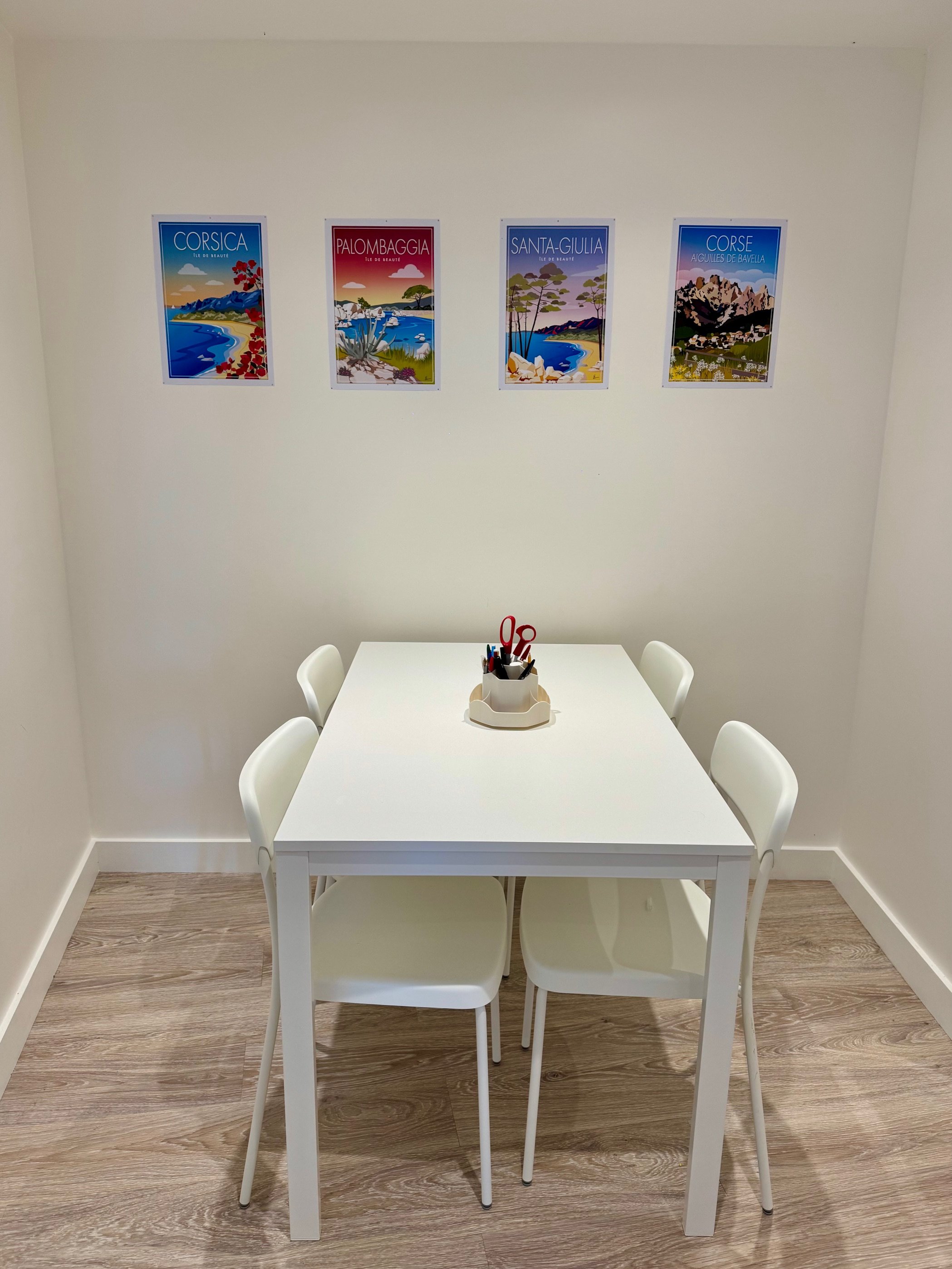 Une petite salle avec une table blanche entourée de quatre chaises blanches, un support pour les fournitures de bureau au centre de la table, et quatre posters colorés représentant différentes régions de Corse accrochés au mur blanc.