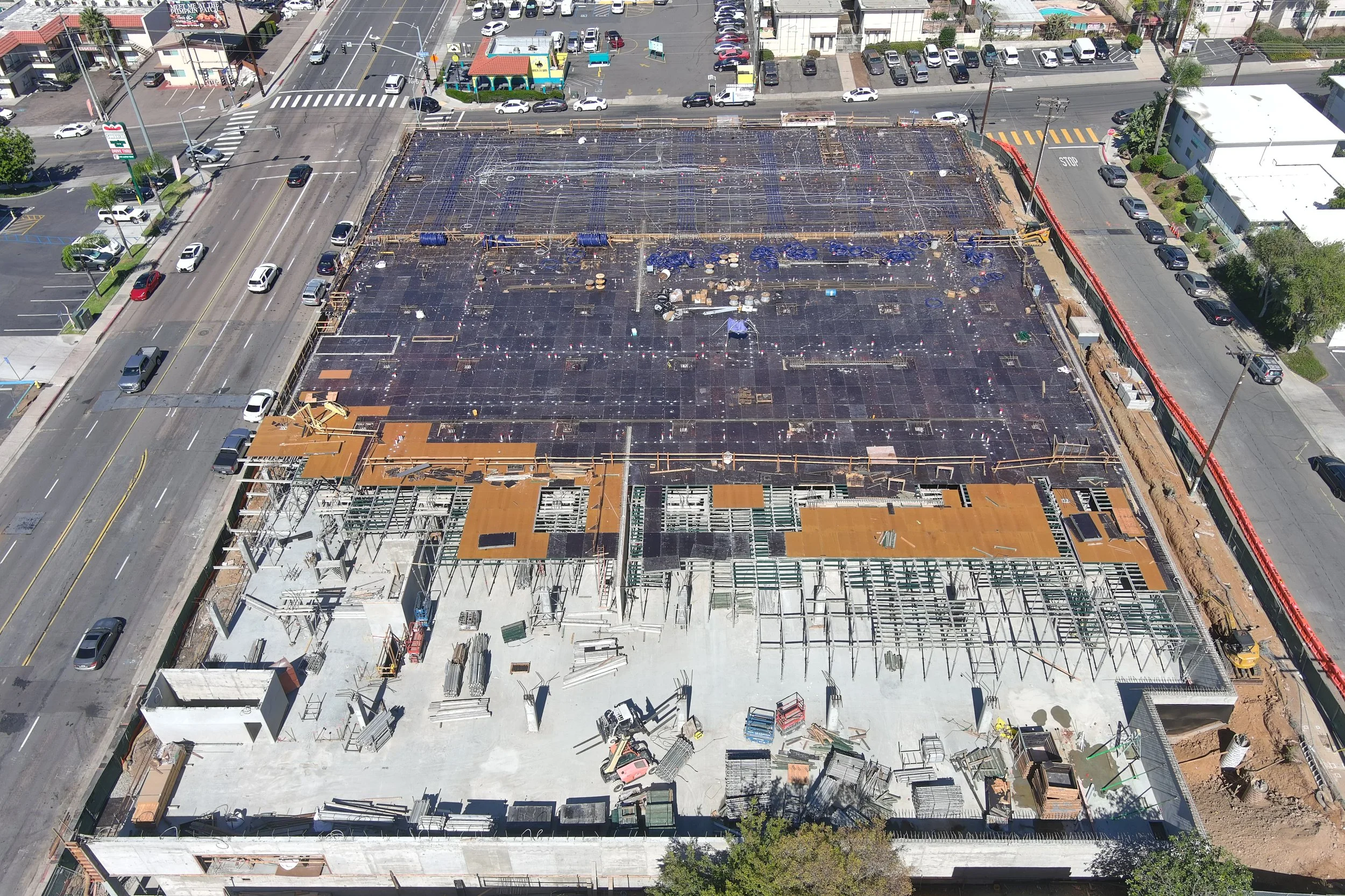 Aerial view of a building under construction, with a steel framework and wooden roof panels, surrounded by busy streets and parked cars.