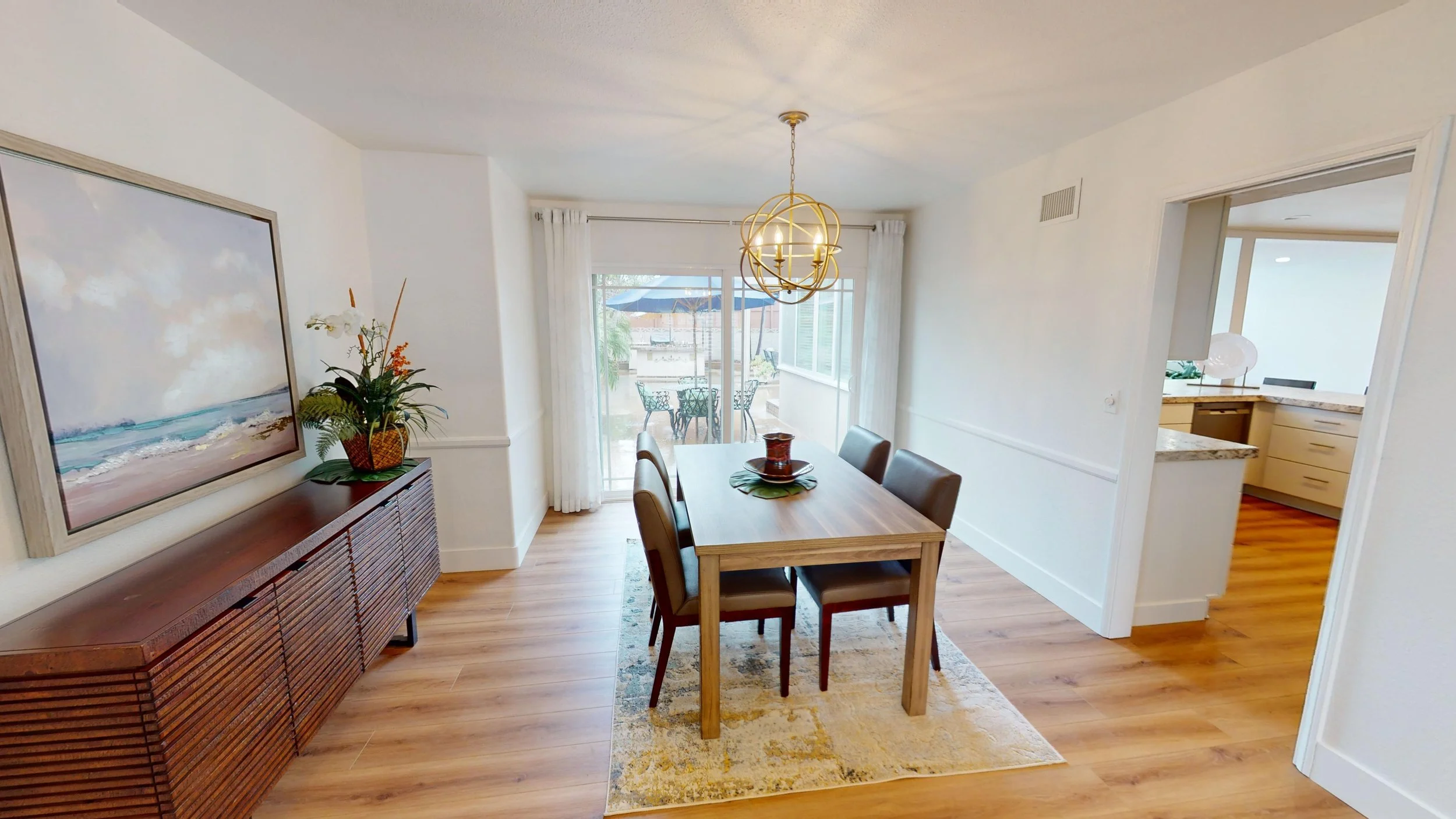 Dining room with wooden table and six chairs, sliding glass door opening to a patio with outdoor furniture, light wood flooring, abstract wall art, and a sideboard with a plant.