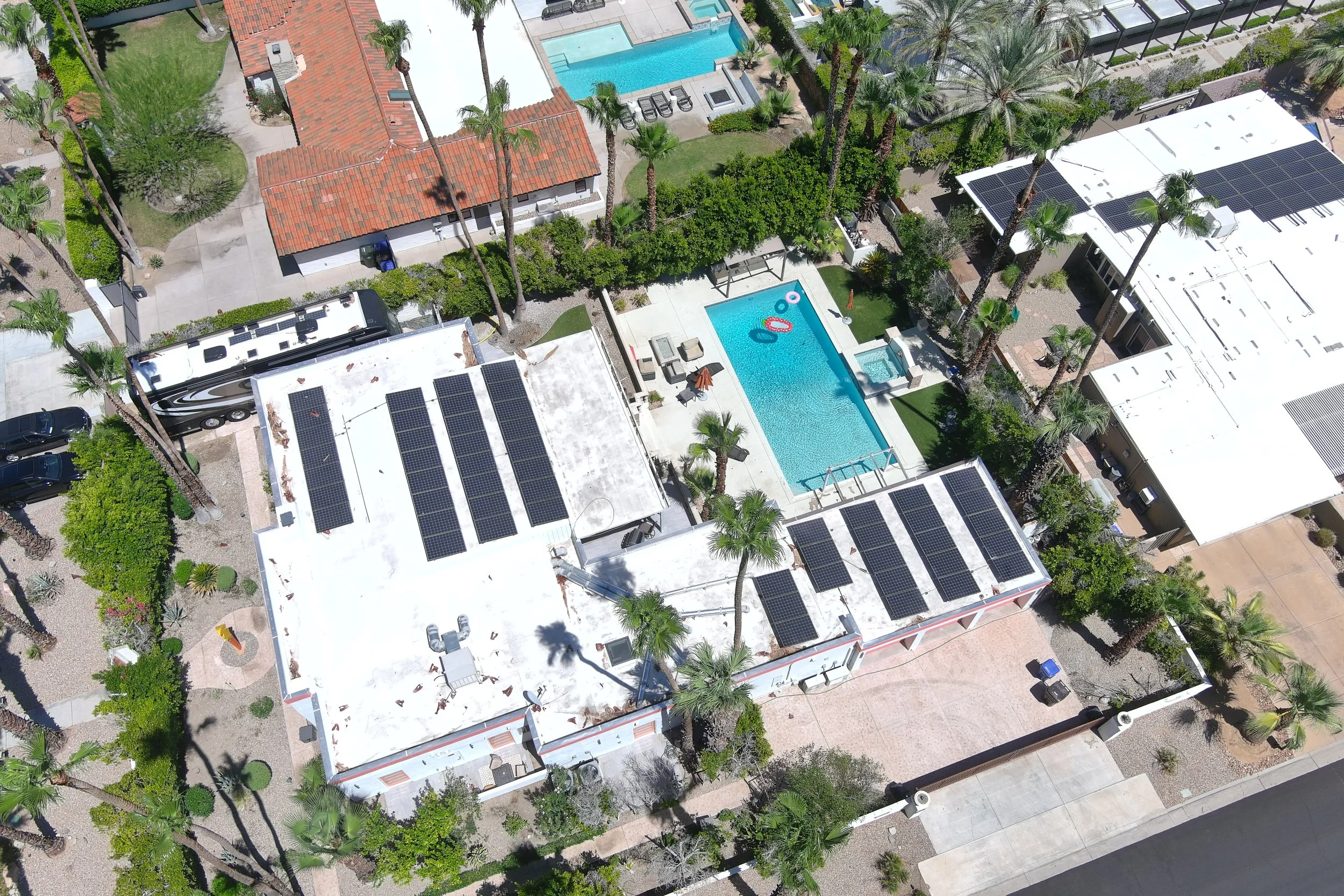 Aerial view of a residential area featuring two houses with solar panels on the roofs, a swimming pool with lounge chairs and inflatables, a separate smaller pool, and a RV parked nearby, surrounded by palm trees and other greenery.