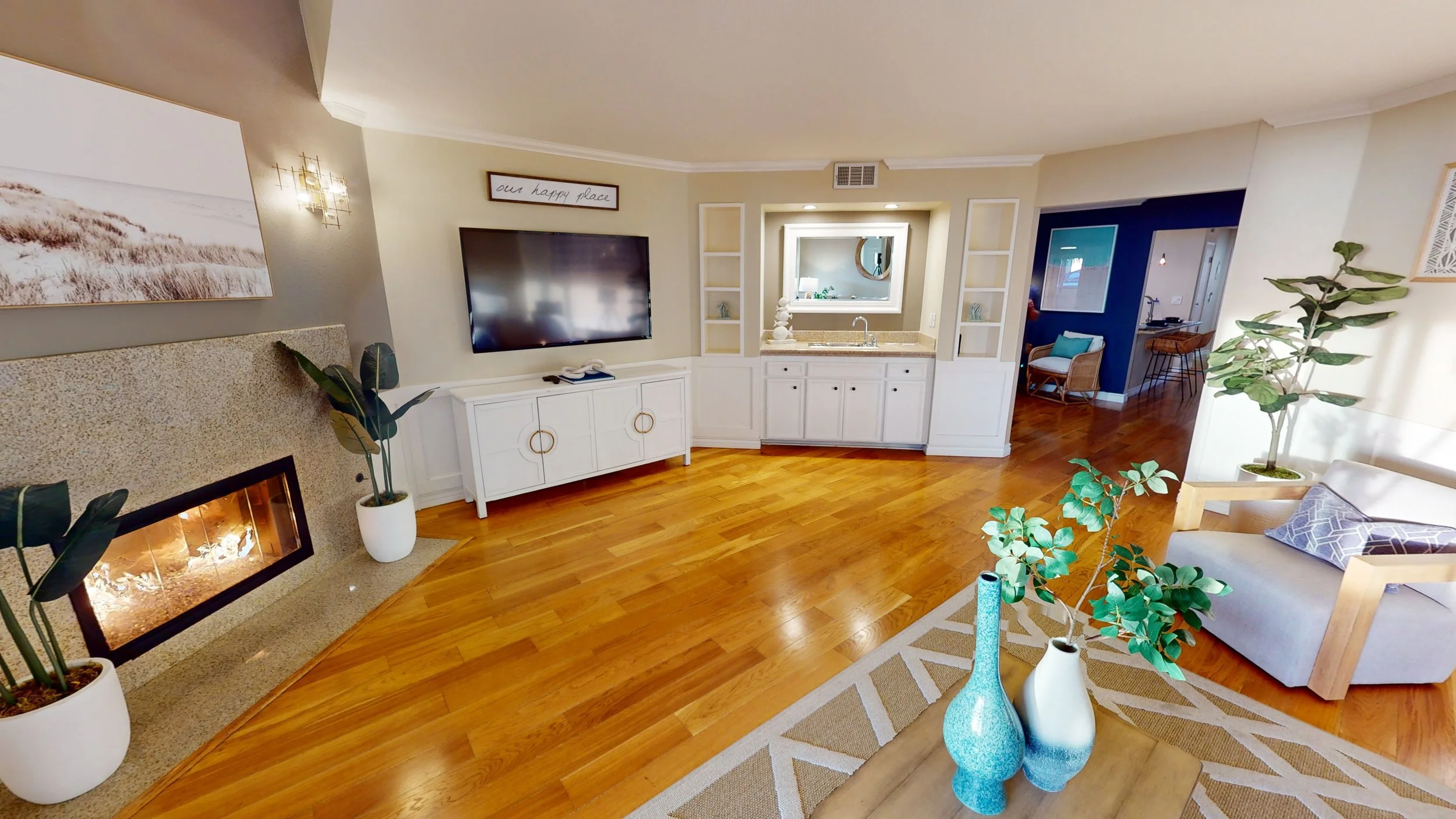 Living room with wooden floor, fireplace, wall-mounted TV, white entertainment console, potted plants, armchair, and decorative vases.