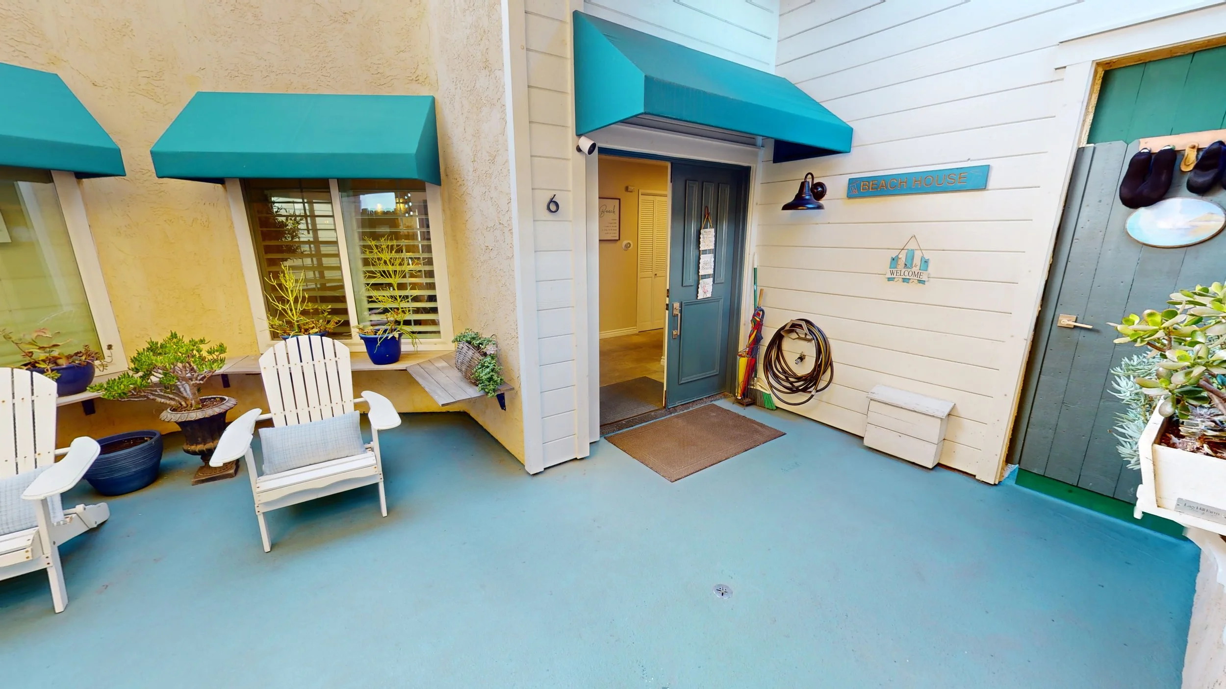 Outdoor entrance to a beach house with white siding, a blue awning over the door, and decorative signs on the wall. There are two white chairs, potted plants, and hanging shoes on the wall.